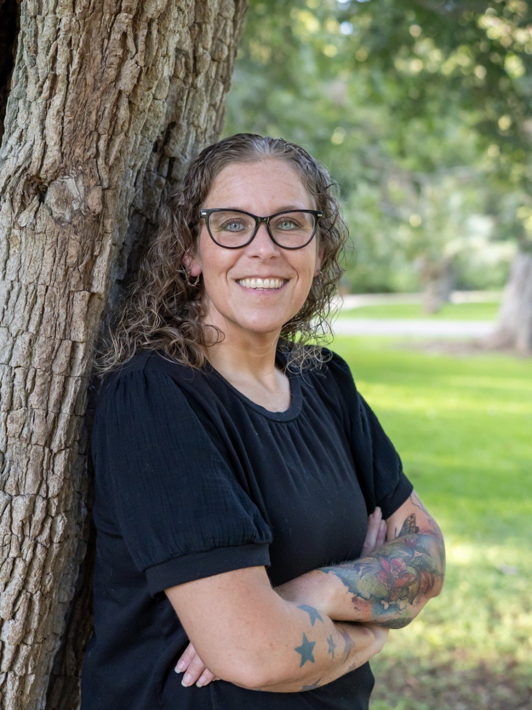 A woman with curly hair and glasses leaning against a tree in a park, smiling at the camera.