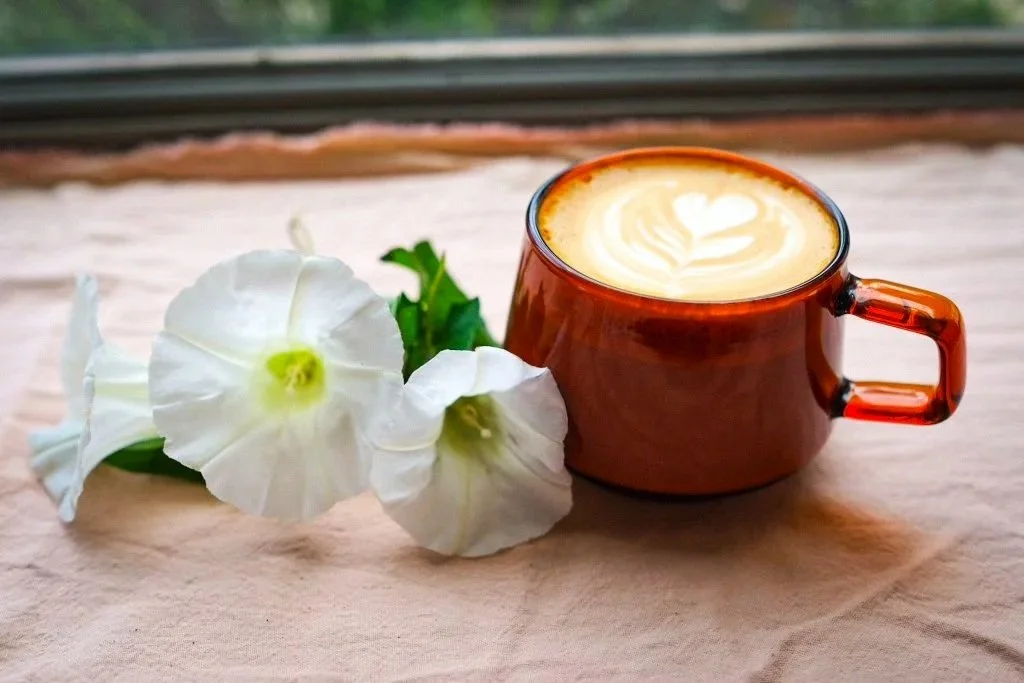 A brown ceramic mug filled with latte featuring a heart-shaped latte art, placed on a light fabric surface alongside white morning glory flowers with green stems.