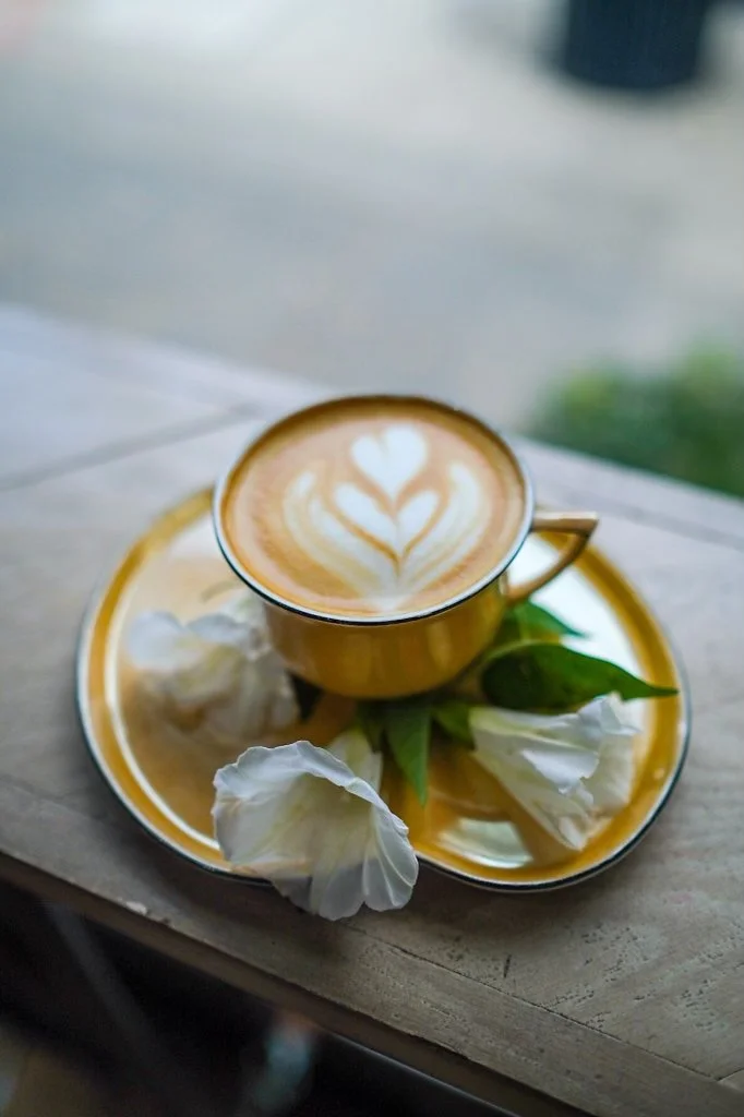 A cup of coffee with latte art on a yellow saucer, garnished with white flowers and green leaves, on a wooden surface.