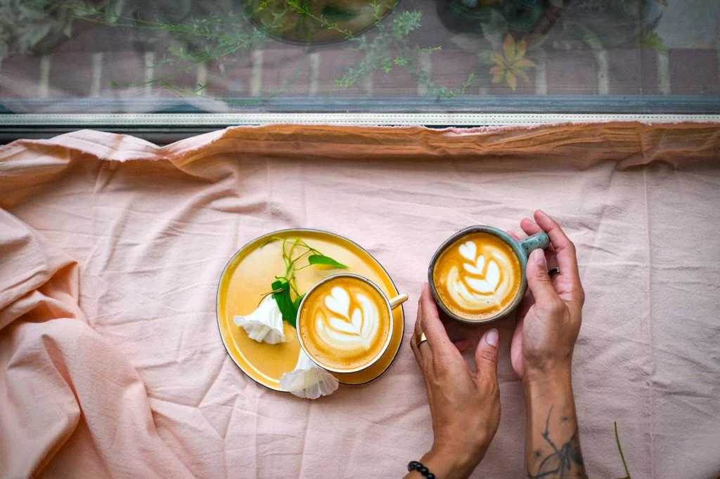 Two cups of latte with heart-shaped latte art, one on a yellow saucer and the other held in a person’s hands, on a pink cloth beside a window with some greenery outside.