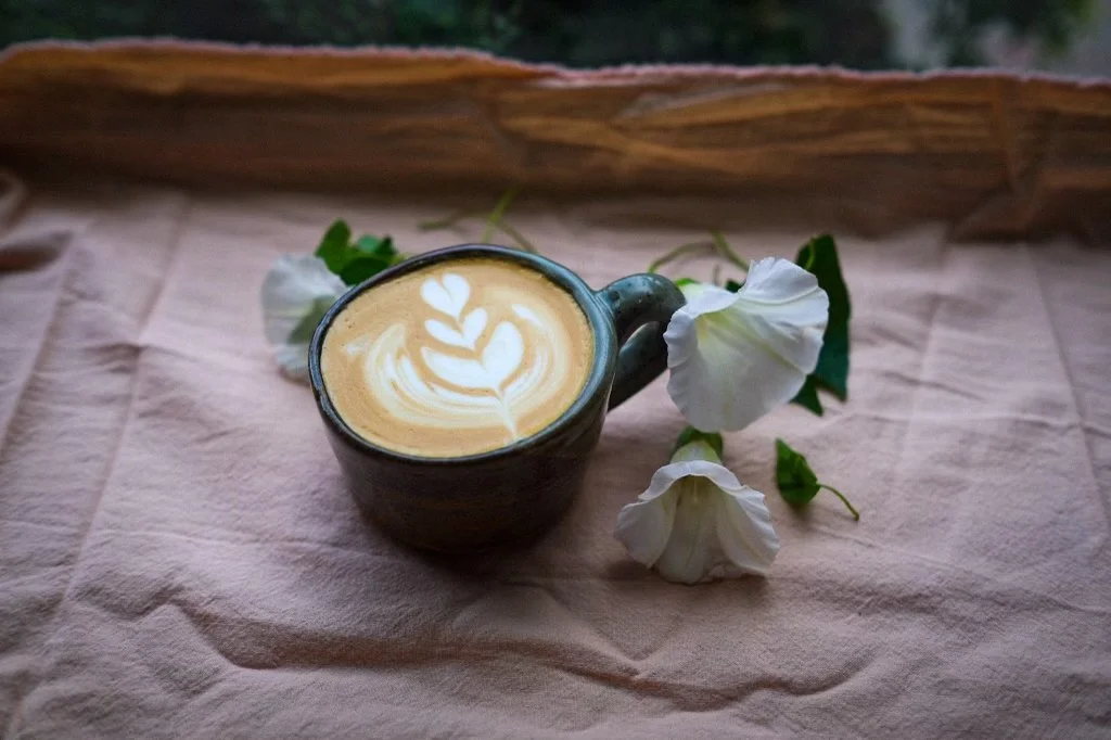 A cup of coffee with latte art on a pink cloth, surrounded by white flowers.