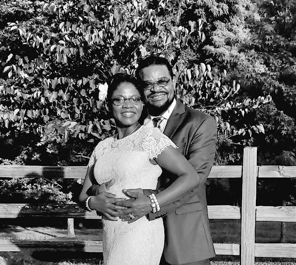 A smiling couple dressed in formal attire stands in front of a wooden fence and leafy trees, embracing each other outdoors.