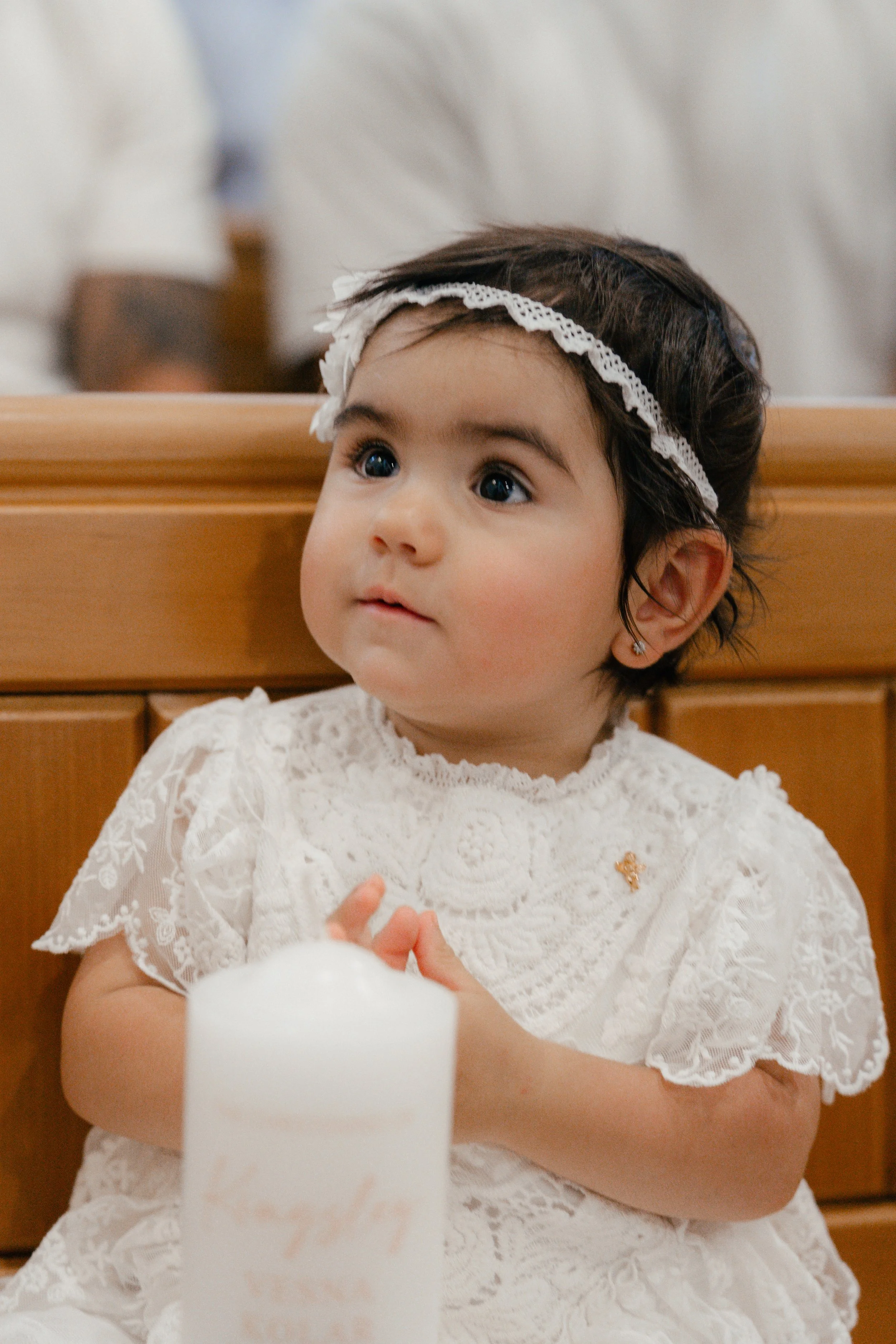 A young girl with dark hair, wearing a white lace dress and a headband, sitting in church, holding her hands together, and looking upward with a curious expression.