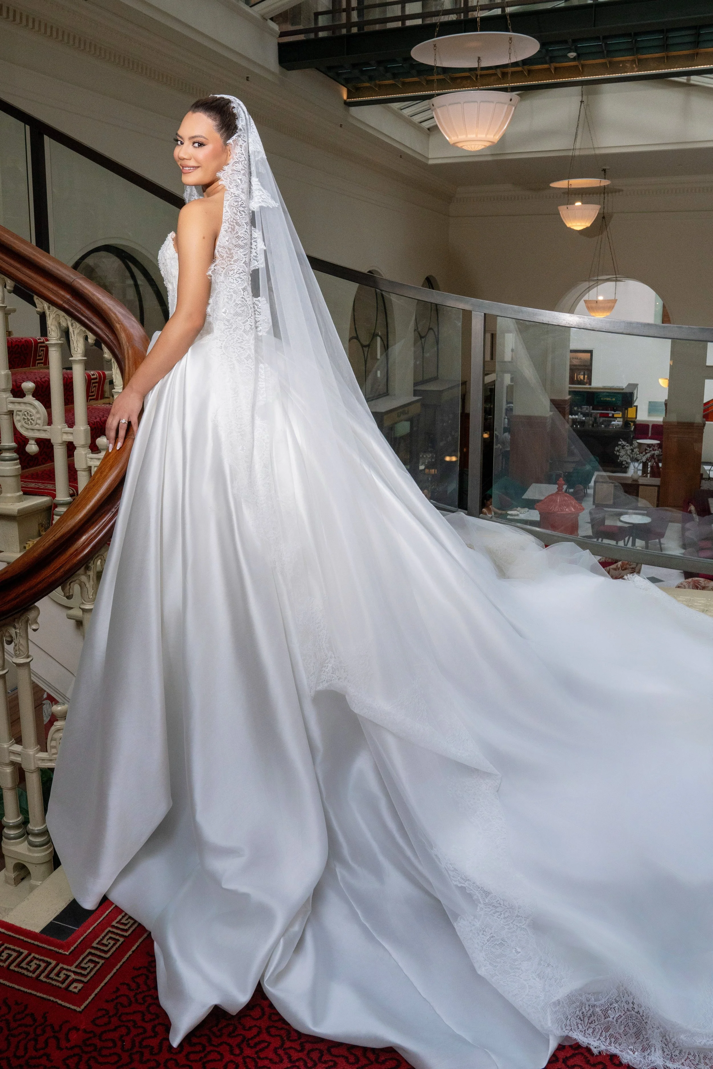 A bride wearing a white wedding gown with a long train and lace veil, smiling on a staircase inside a decorated venue.
