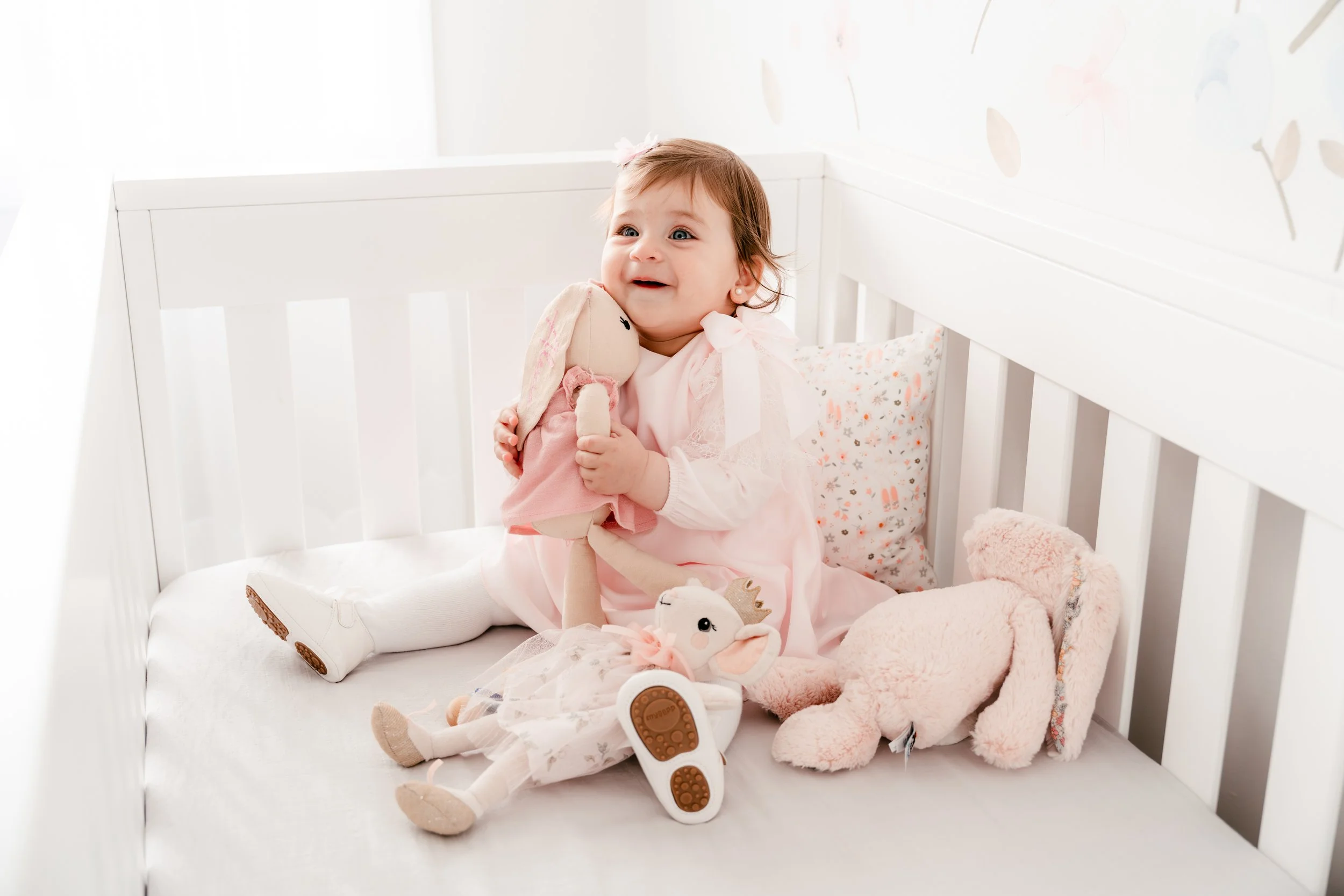 A young girl sitting in a white crib, holding a plush bunny toy, surrounded by other stuffed animals, with light-colored bedding and pillows, in a bright, softly lit nursery.
