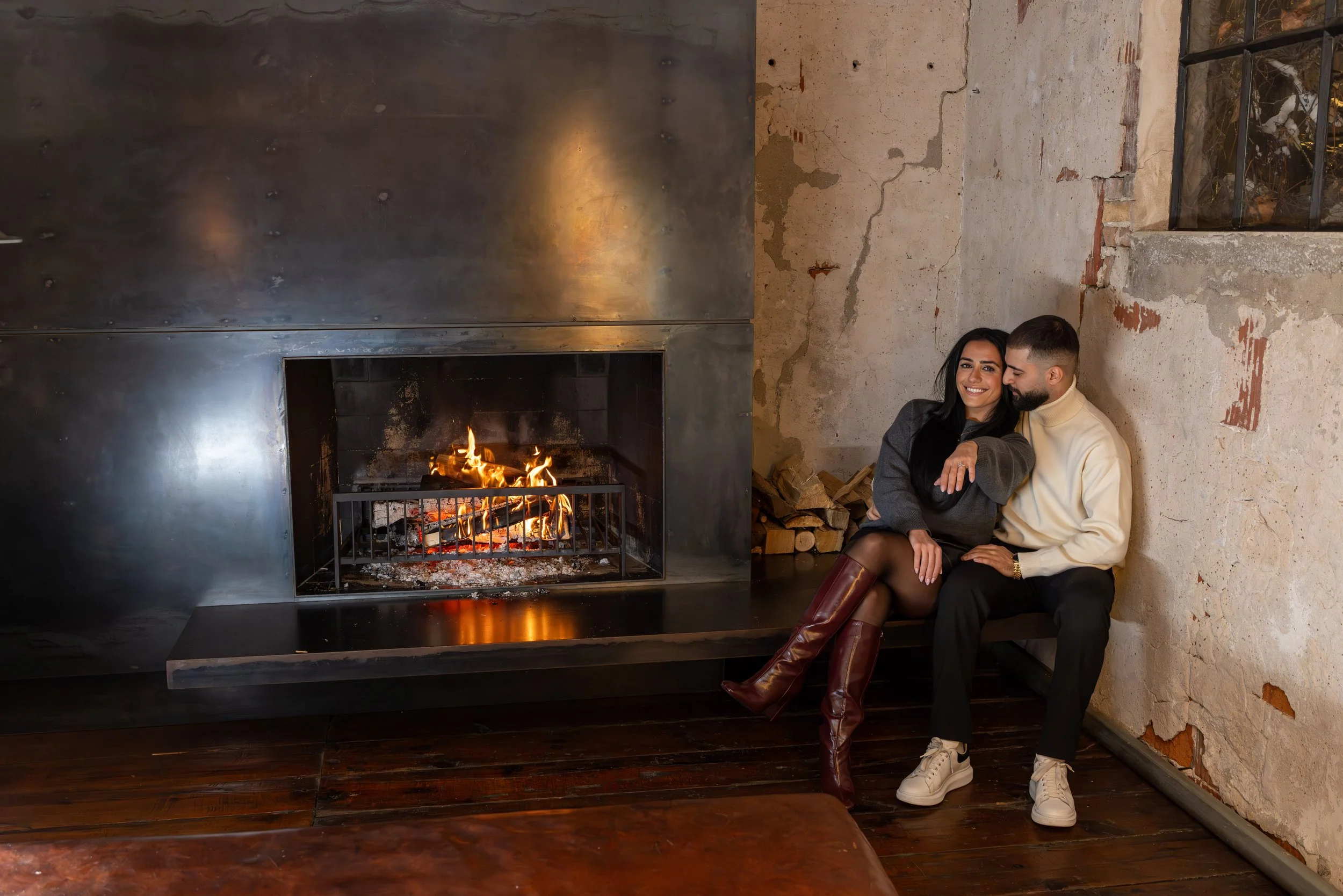 A couple sitting on a bench next to a modern fireplace with a fire burning inside, in a rustic room with peeling plaster walls and a window.
