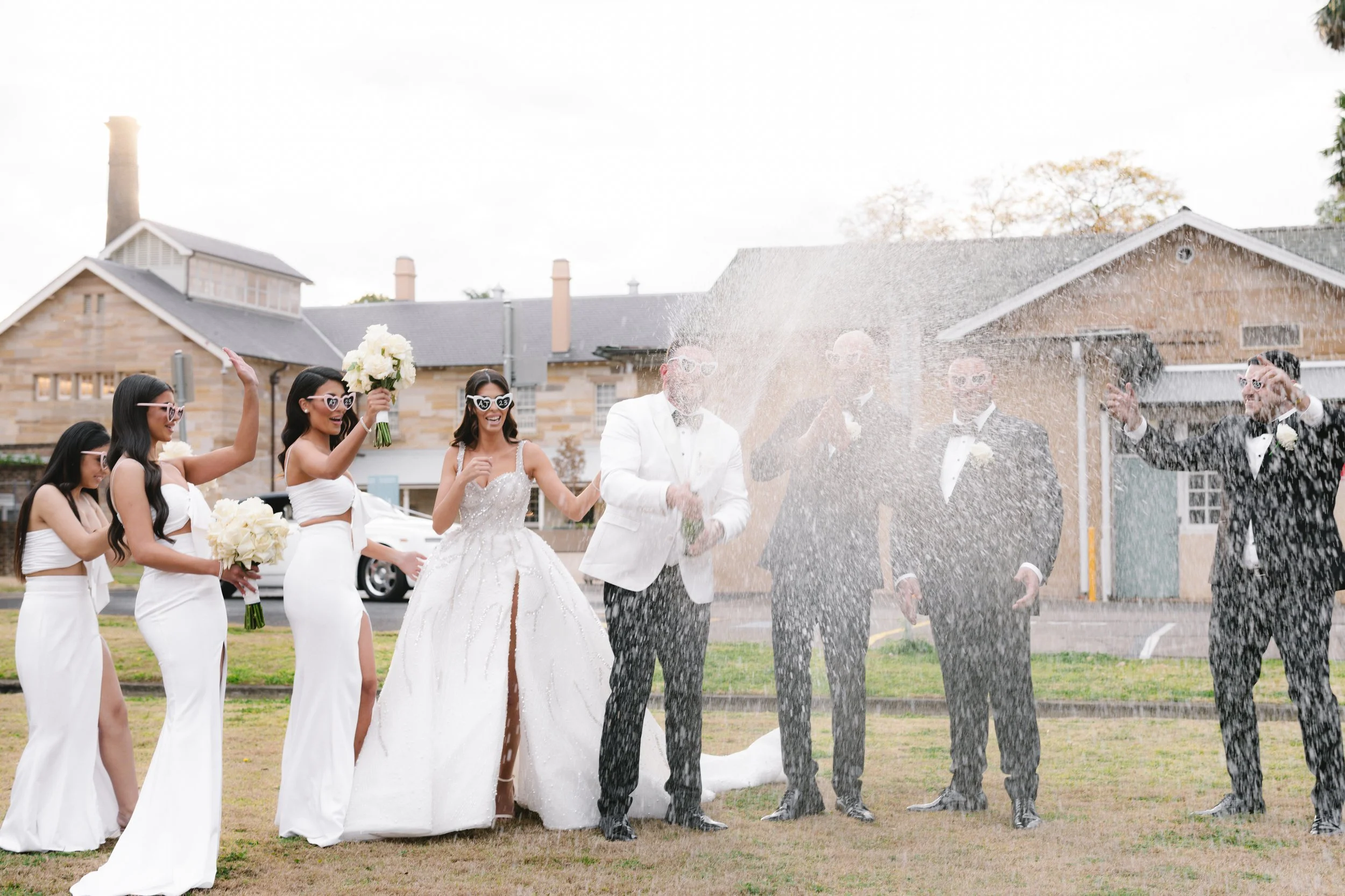 Wedding celebration with bride and groom spraying champagne outdoors, surrounded by bridesmaids and groomsmen.