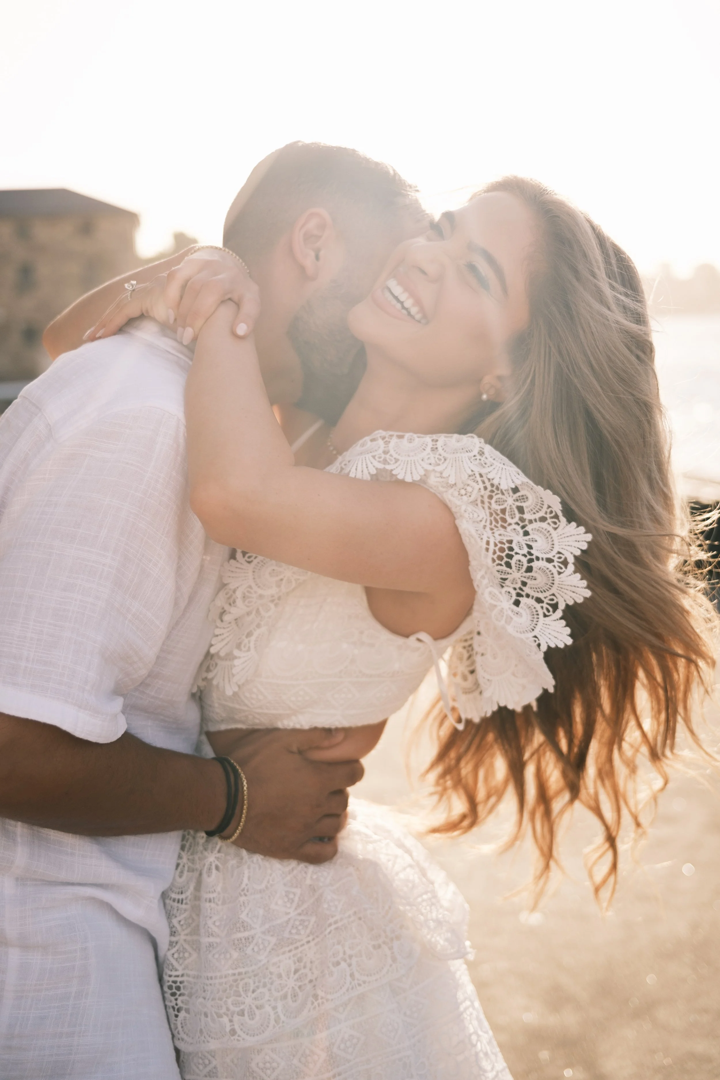 A happy couple embracing outdoors at sunset, with the woman smiling and the man holding her close.