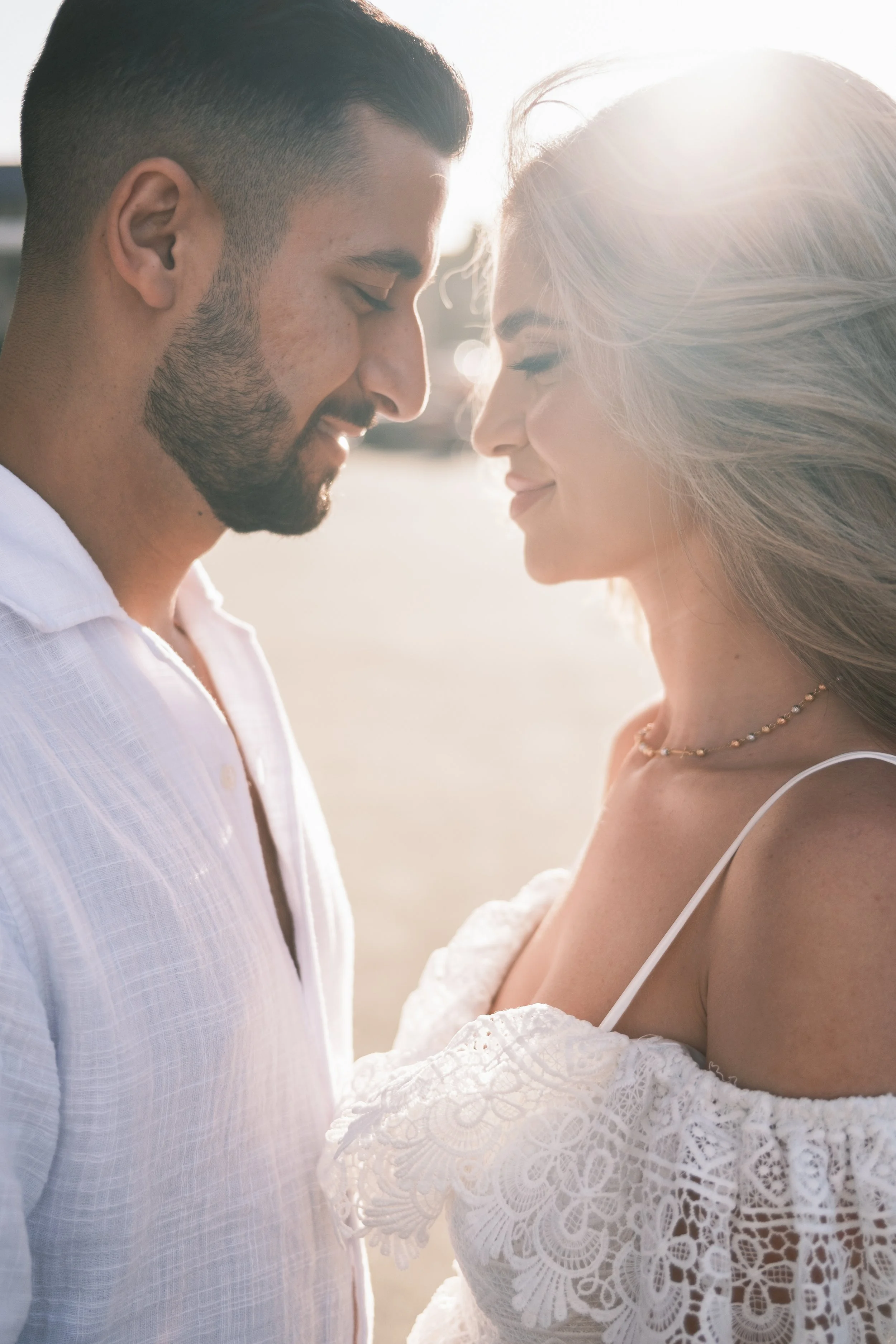 A man and woman with foreheads touching, smiling gently at each other, on a sunny outdoor setting, dressed in white clothing.