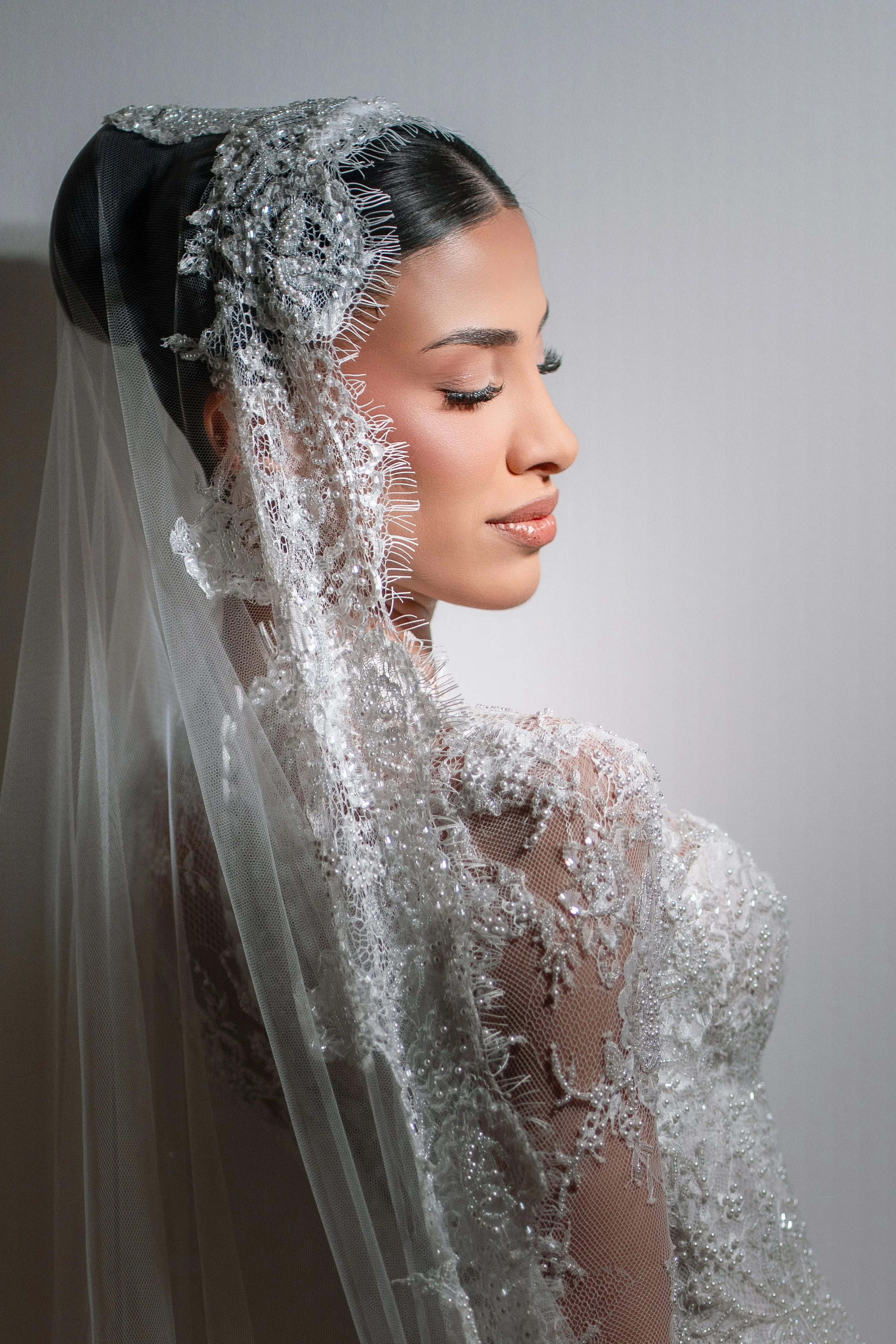 Close-up of a woman in an elegant, embroidered wedding dress with lace details, wearing a jeweled headpiece and veil, with her eyes closed and a peaceful expression.