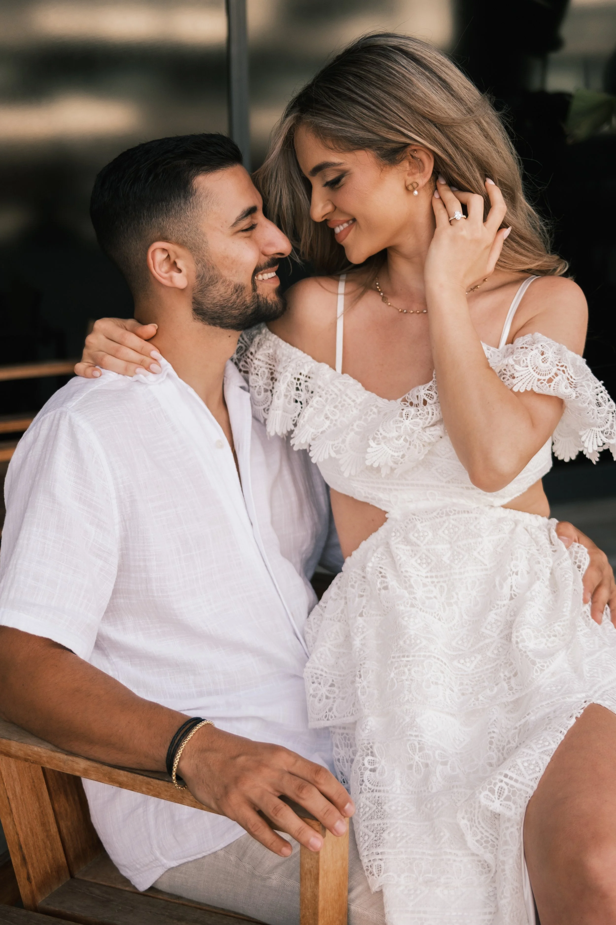 A man and woman sitting close together, smiling and looking into each other's eyes, with the woman touching her ear and wearing a white lace dress, and the man wearing a white shirt.