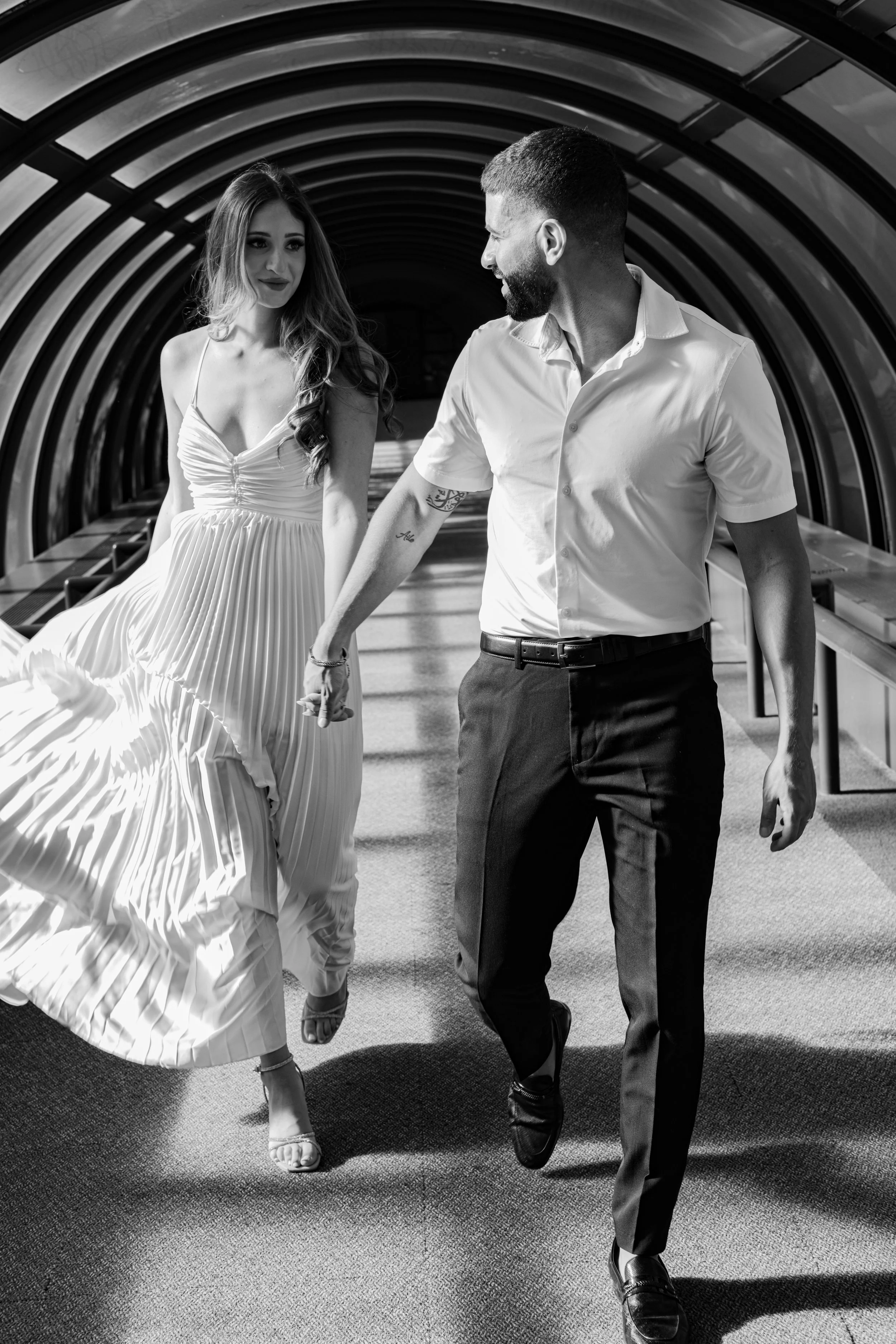 A man and a woman walking hand in hand through an arched, tunnel-like structure, with the woman wearing a pleated dress and high heels and the man dressed in a button-up shirt and slacks, both smiling.