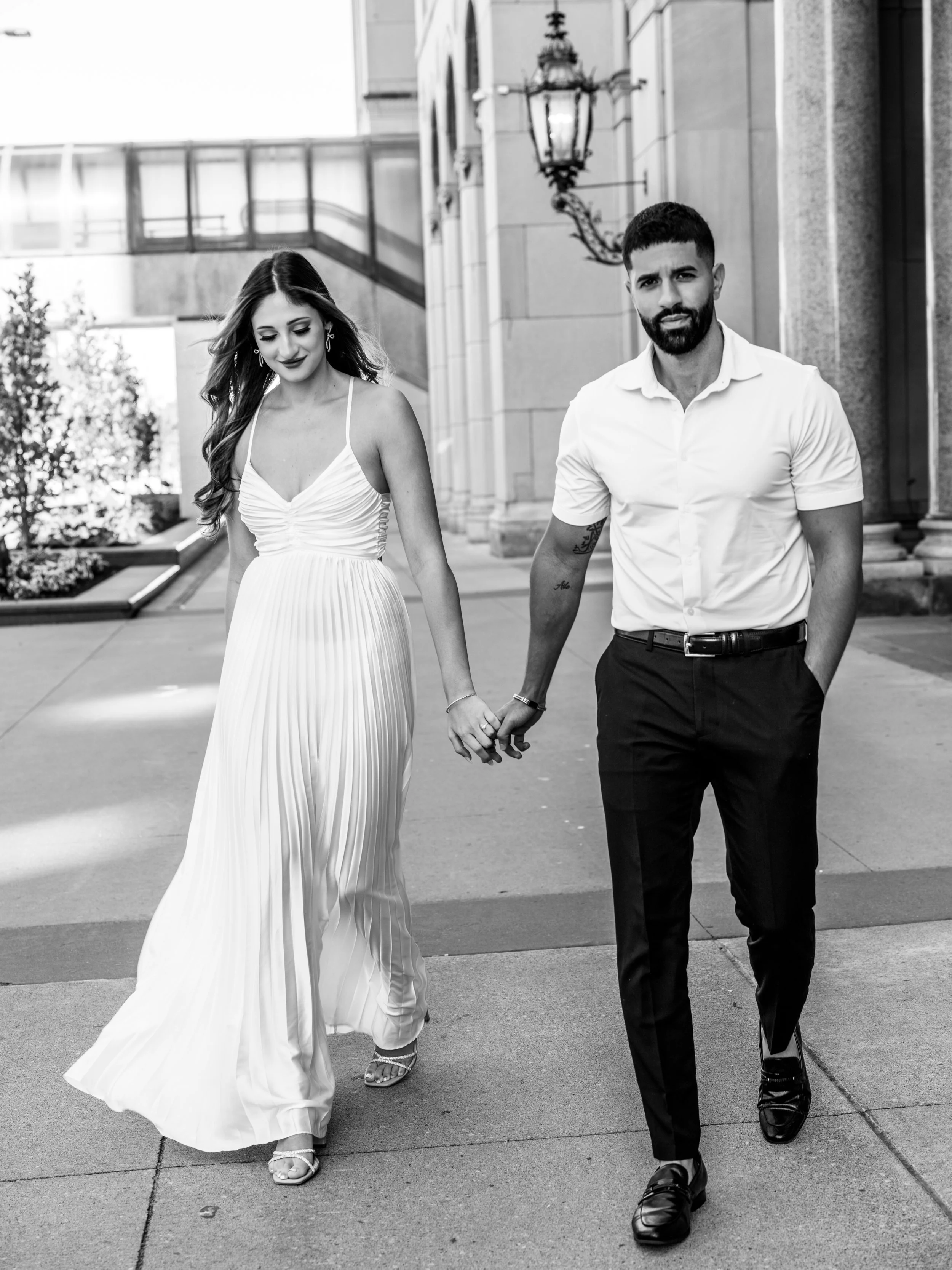 A young couple holding hands is walking outdoors on a city sidewalk. The woman is wearing a flowing, pleated white dress and high heels, with long hair and earrings. The man is dressed in a short-sleeved white shirt, dark pants, and loafers, with a b