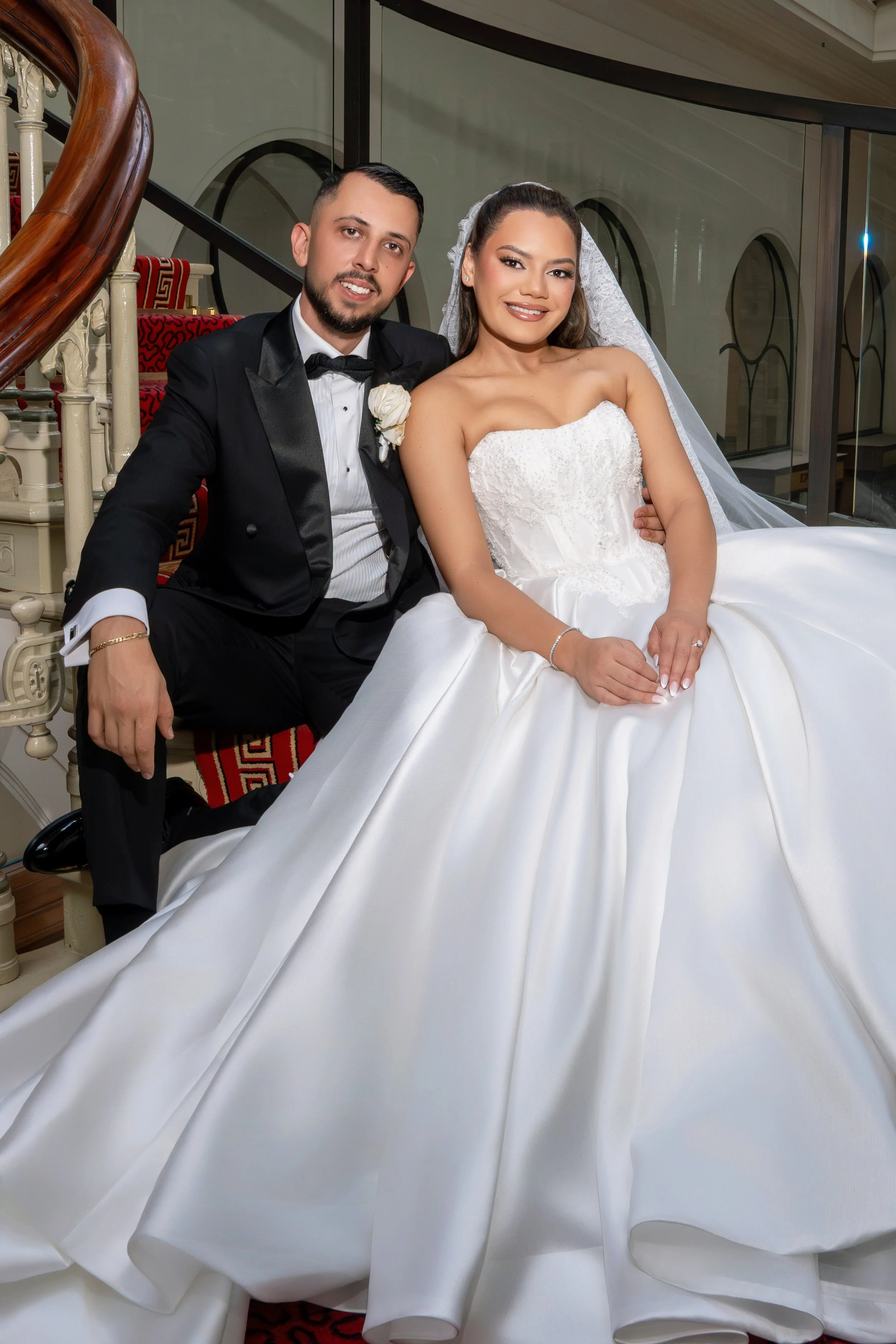 A bride and groom sitting together on a staircase at a wedding. The bride is wearing a white wedding gown with lace details and a veil, and the groom is in a black tuxedo with a bow tie.