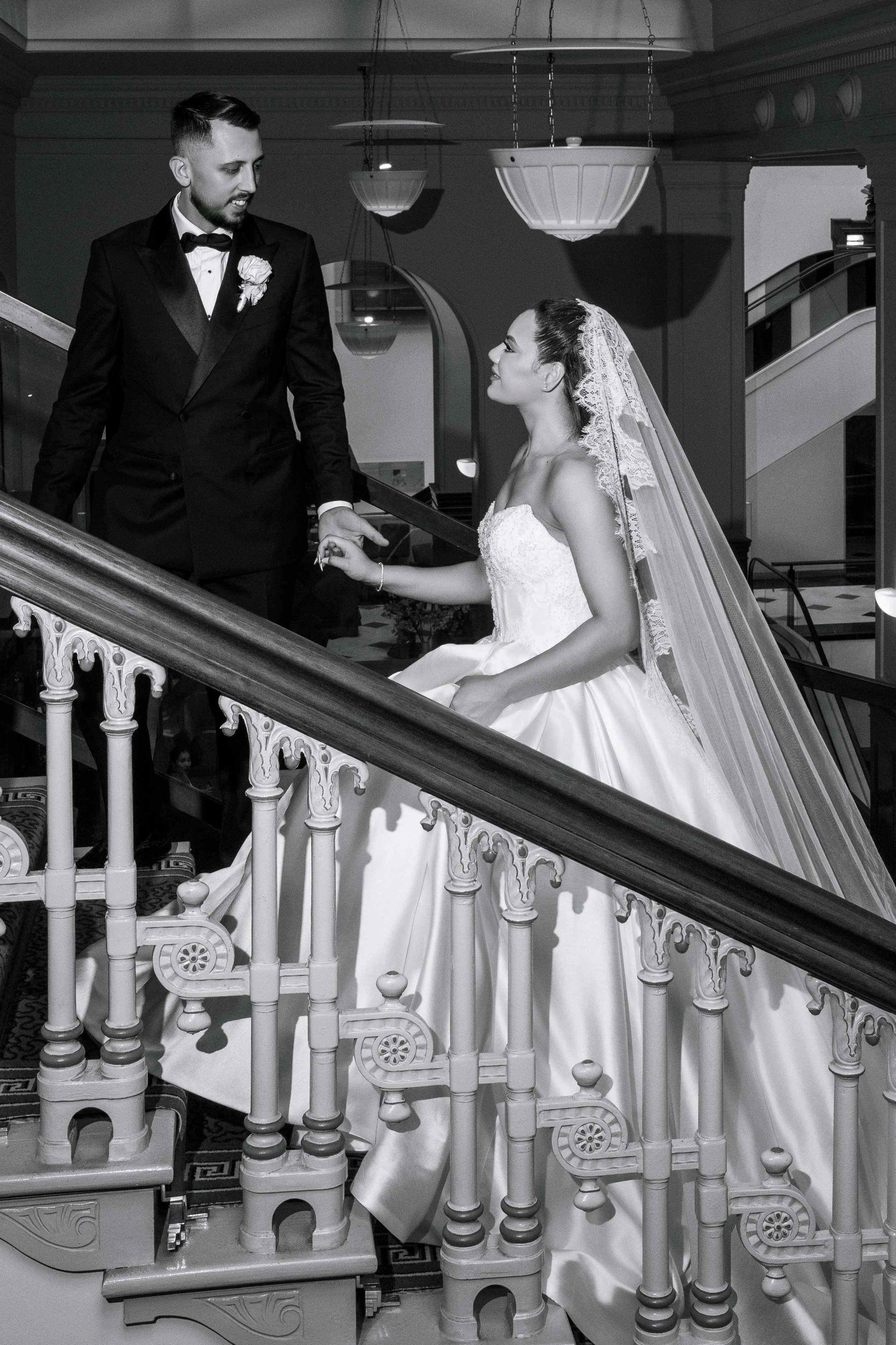 A black and white photo of a bride and groom on a staircase inside a building, with the groom holding the bride's hand and looking at her.