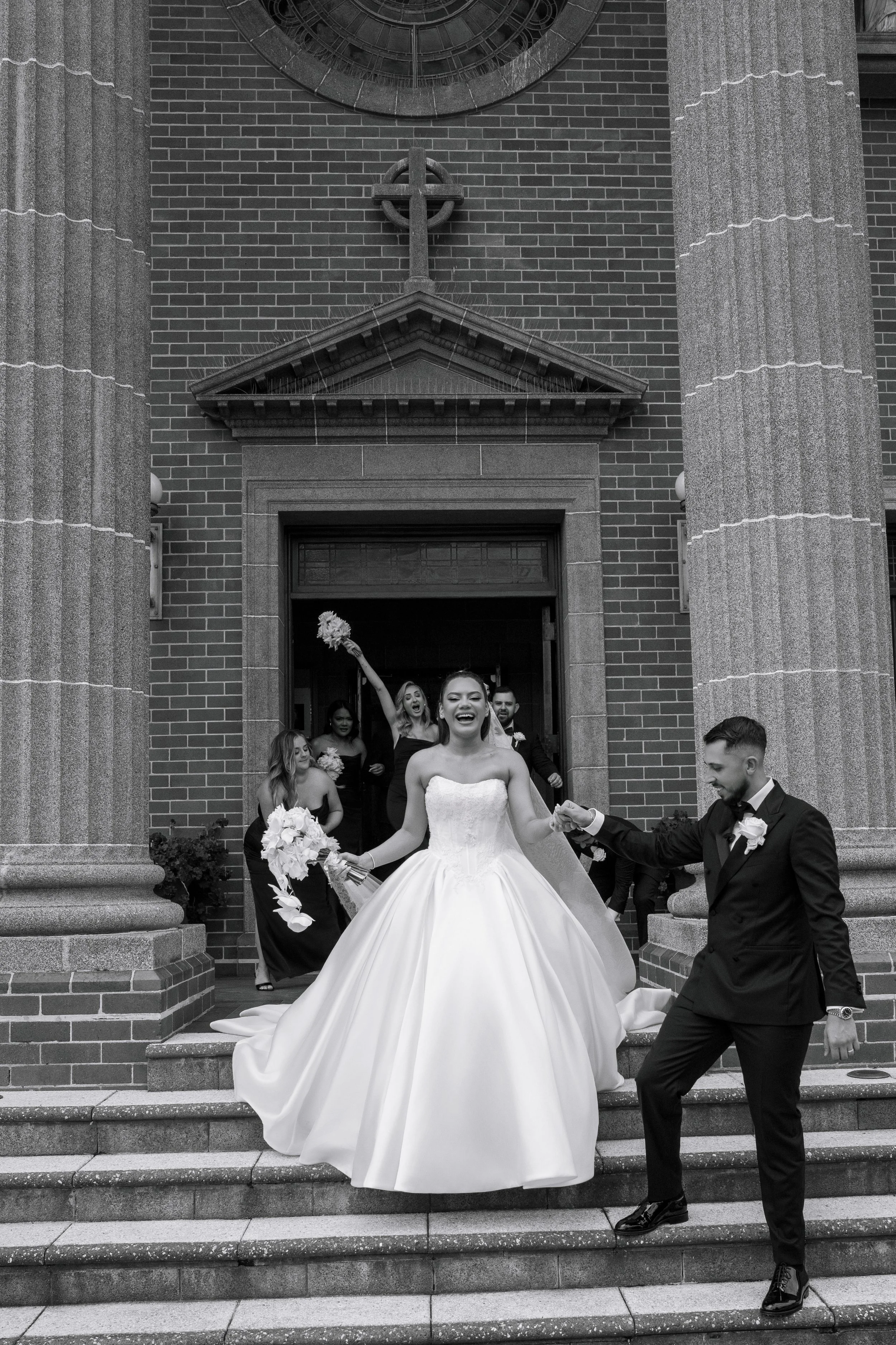 A joyful bride in a wedding gown and a groom in a tuxedo smiling and holding hands on wedding steps outside a church, with wedding party members celebrating behind them.