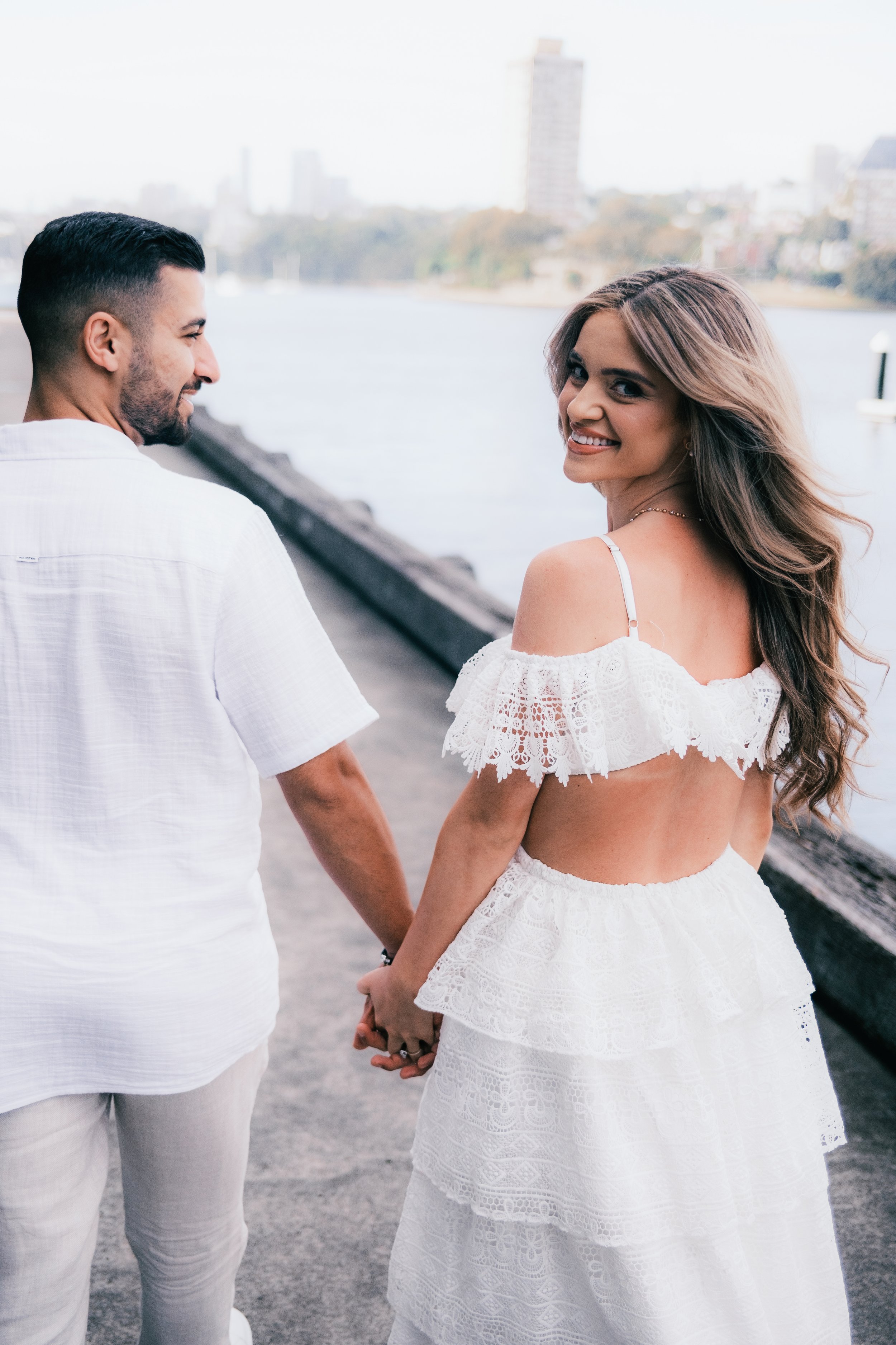 A smiling woman in a white lace dress holding hands with a man in a white shirt by a riverside with city buildings in the background.