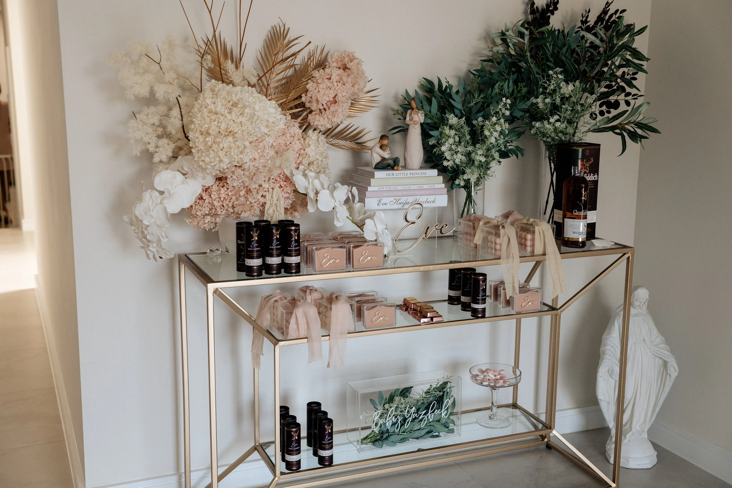 A decorative display with flowers, books, and personalized gifts on a glass and gold console table, with a sculpture of a person on the floor next to it.