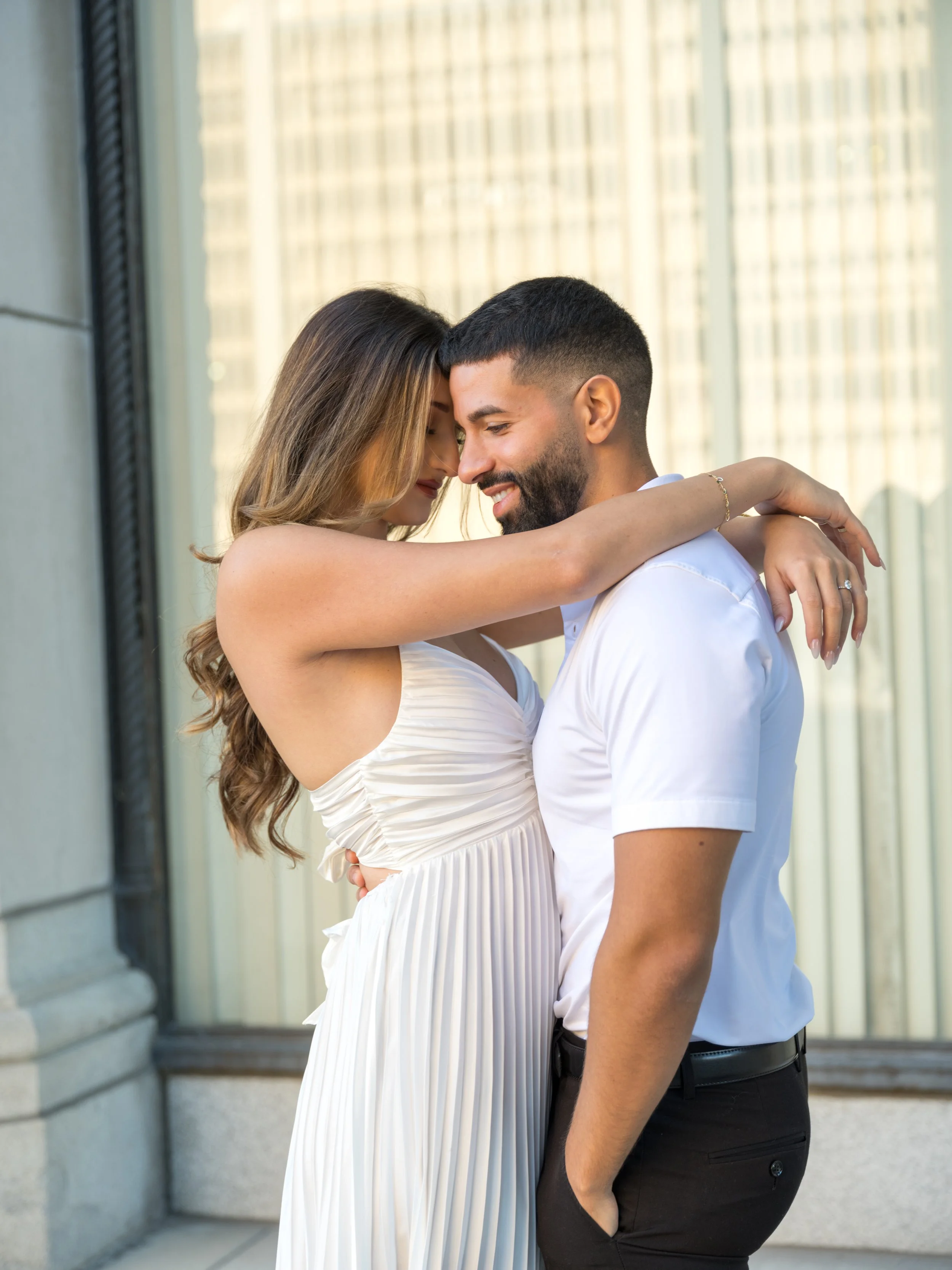 A couple embraces outside a building with large windows, with the woman wearing a sleeveless white dress and the man in a white shirt and dark pants, their foreheads touching and smiling.