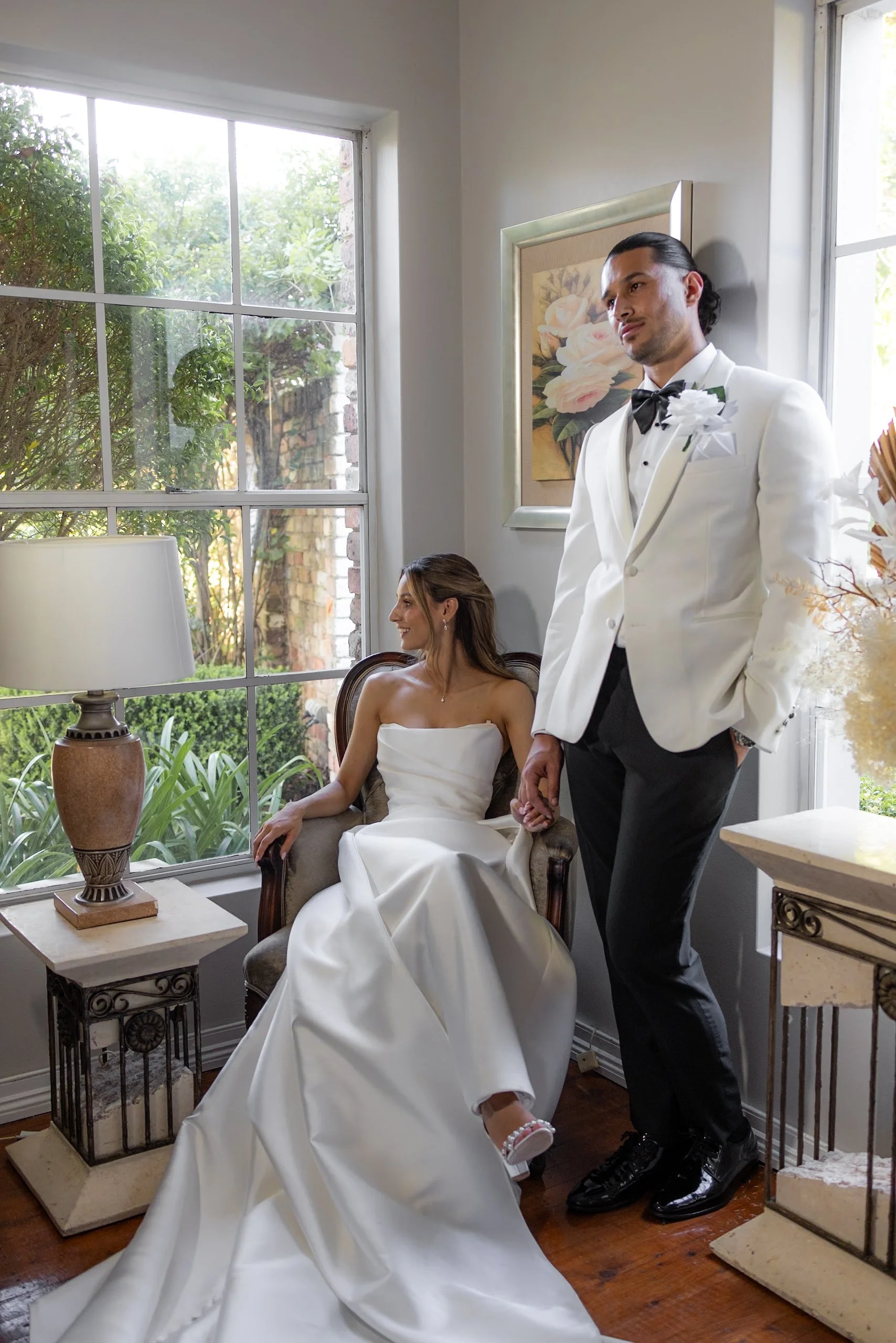 A bride sitting on a chair inside a room, wearing a wedding dress, smiling, with a groom standing beside her wearing a white tuxedo and black bow tie, holding her hand. The room has large windows with a view of greenery outside, a table lamp, and a floral painting on the wall.