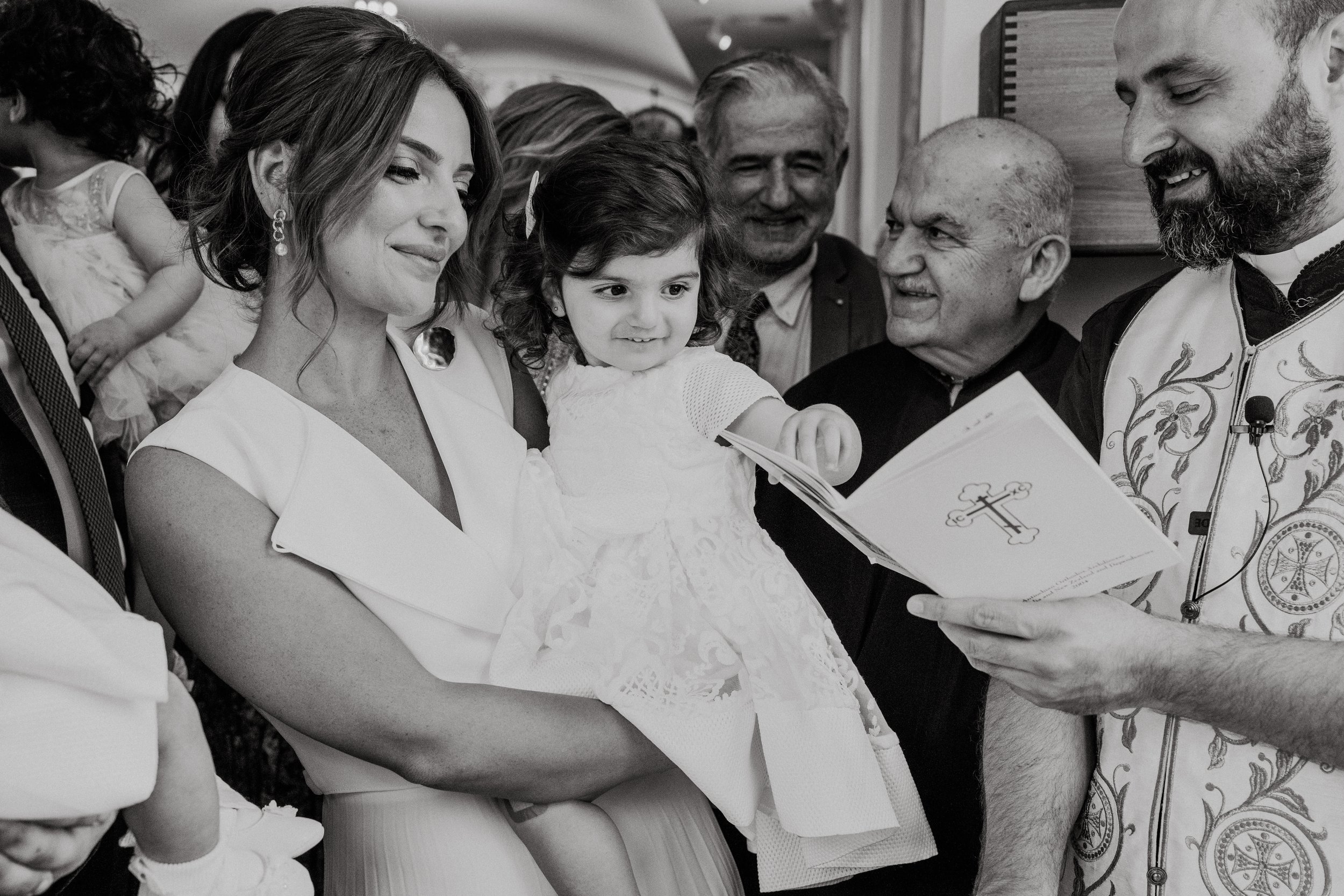 A family at a religious ceremony, with a priest reading from a booklet, and a woman holding a young girl in a white dress looking at a book, surrounded by smiling people.