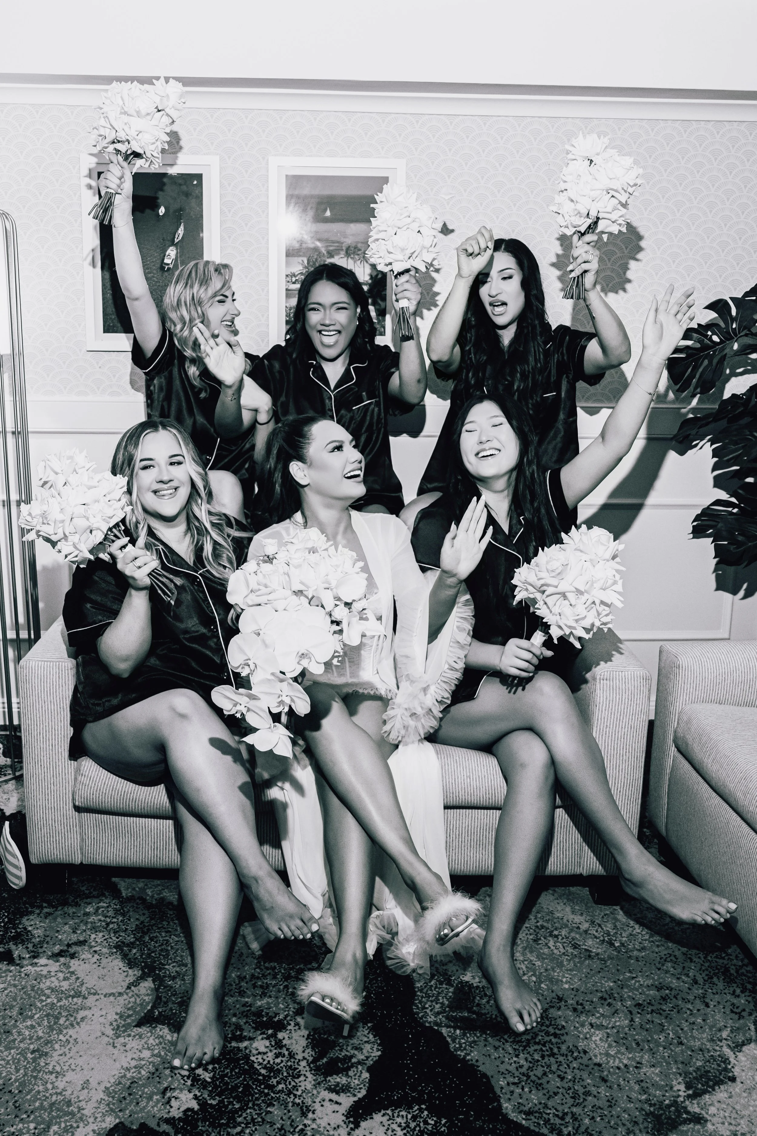 The bride and her bridesmaids in pajamas are celebrating with bouquets of flowers in a living room.