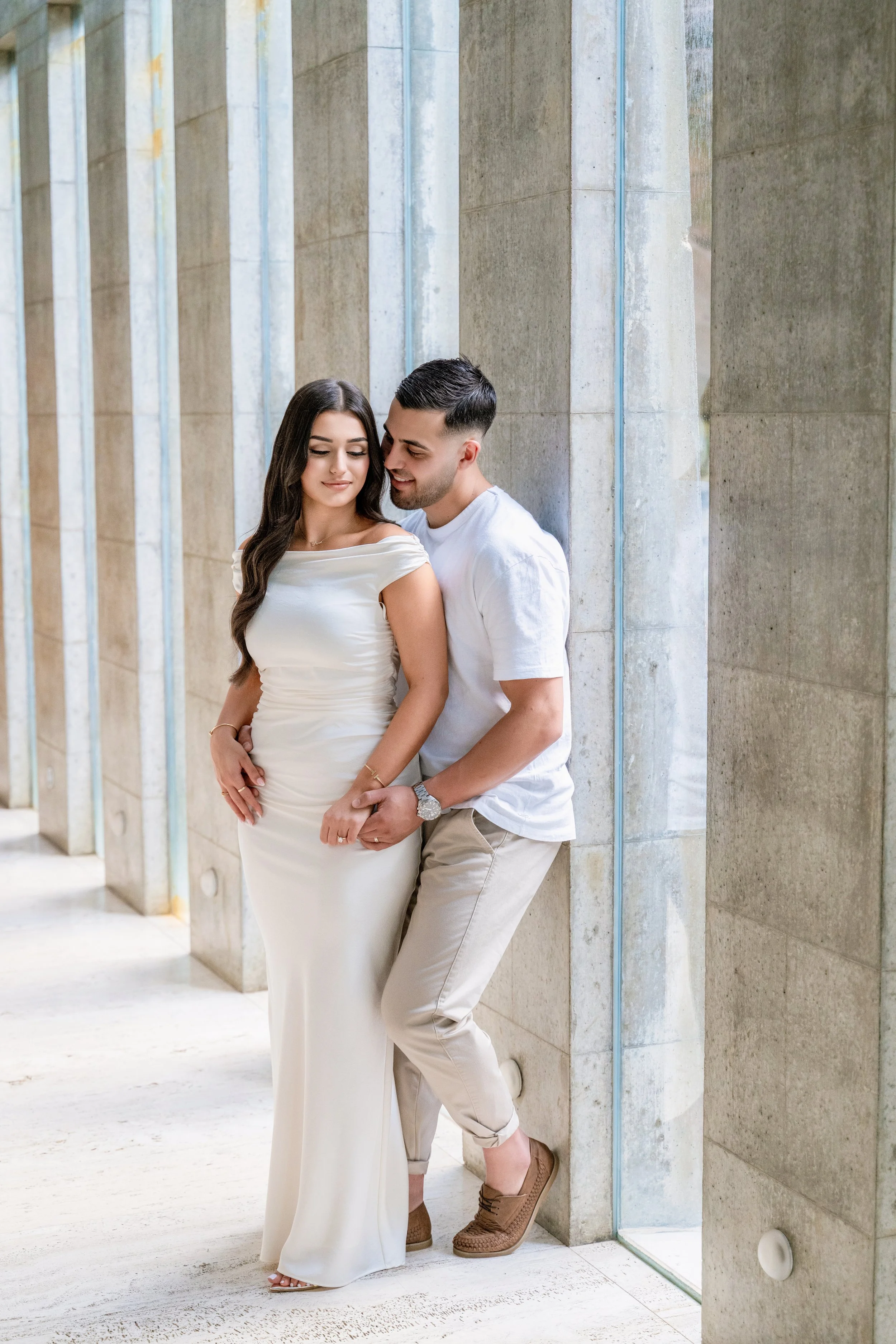 A couple standing close together against a stone wall, with the woman in a white dress and the man in casual clothing, sharing an intimate moment.