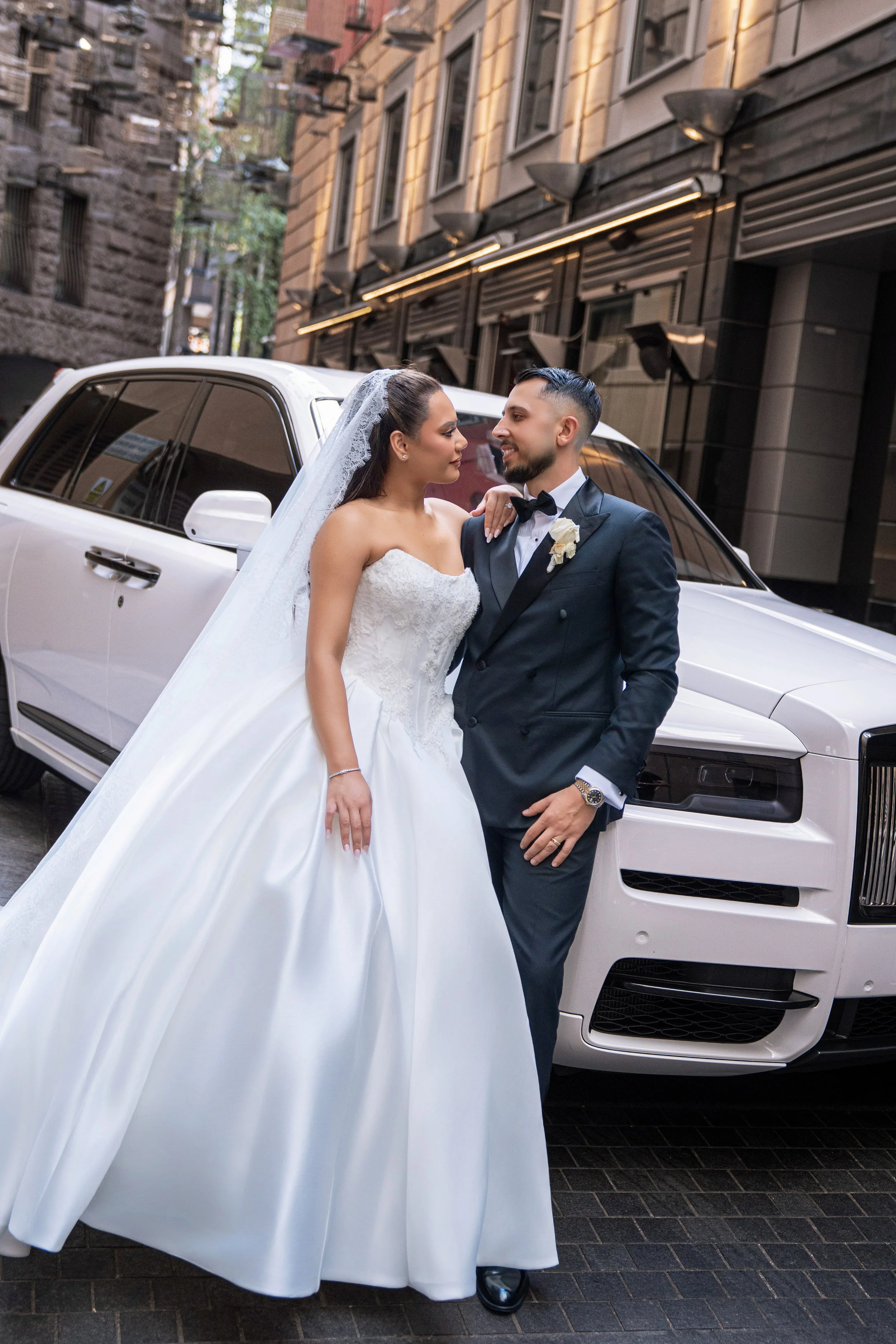 Bride and groom posing together in front of a white car on a city street, dressed in wedding attire, with the bride in a white wedding gown and the groom in a black tuxedo.