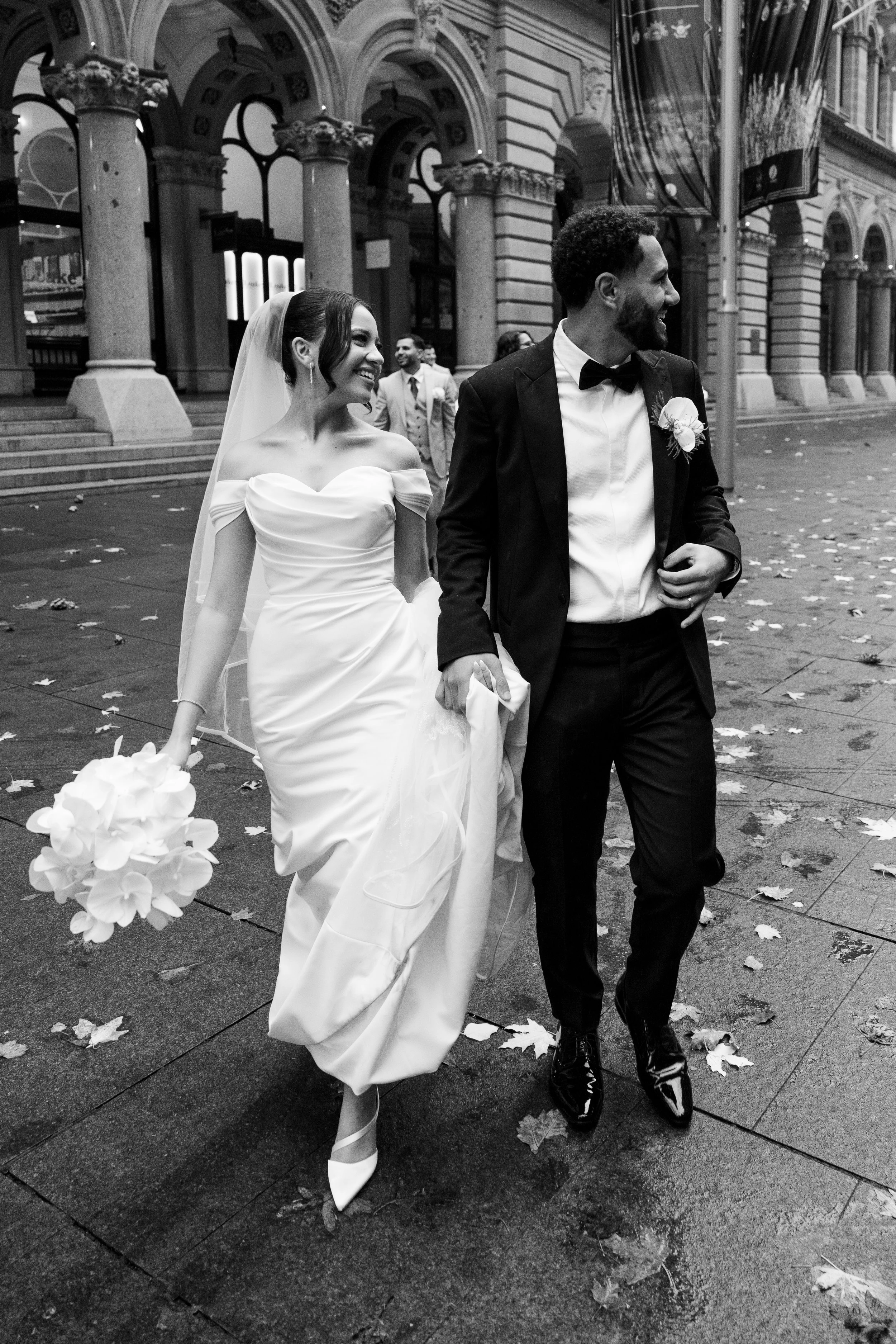 A bride and groom walking on a city sidewalk after their wedding, smiling and holding hands, surrounded by fallen leaves, with historic building architecture in the background.
