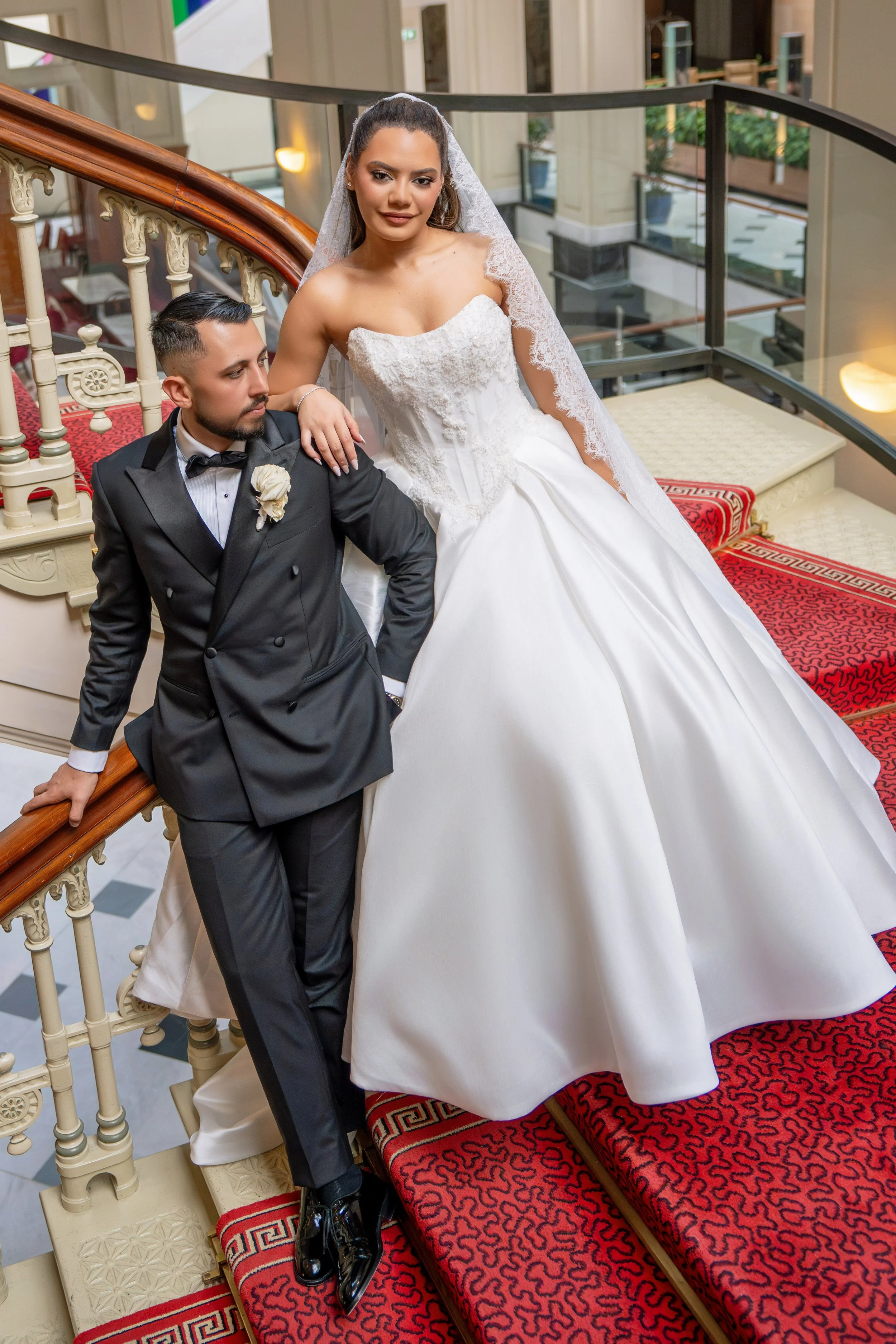 A bride in a white wedding gown and veil, and a groom in a black tuxedo, pose on a staircase at a wedding venue.