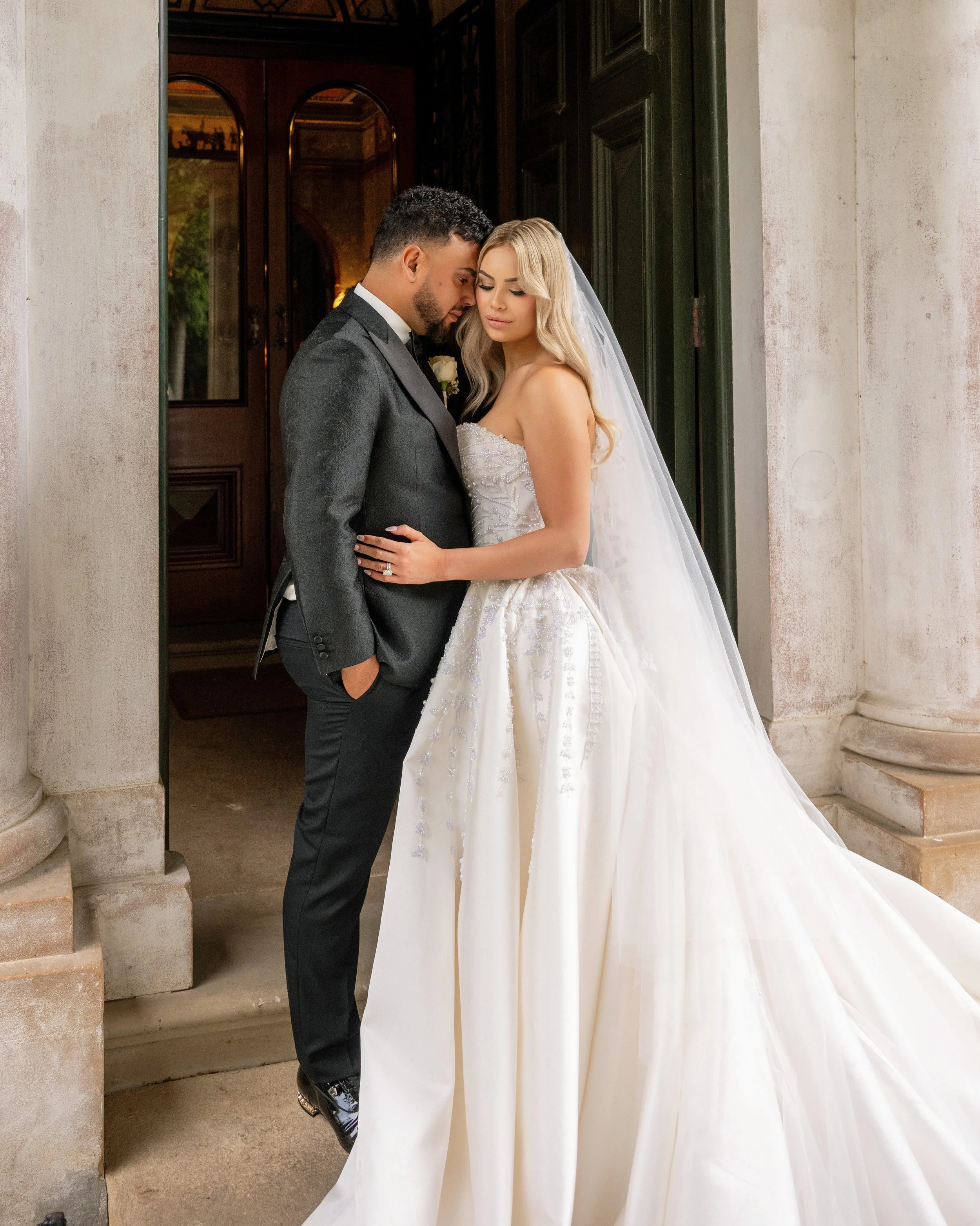 A bride and groom in wedding attire standing close together in front of a building entrance, embracing and looking down.