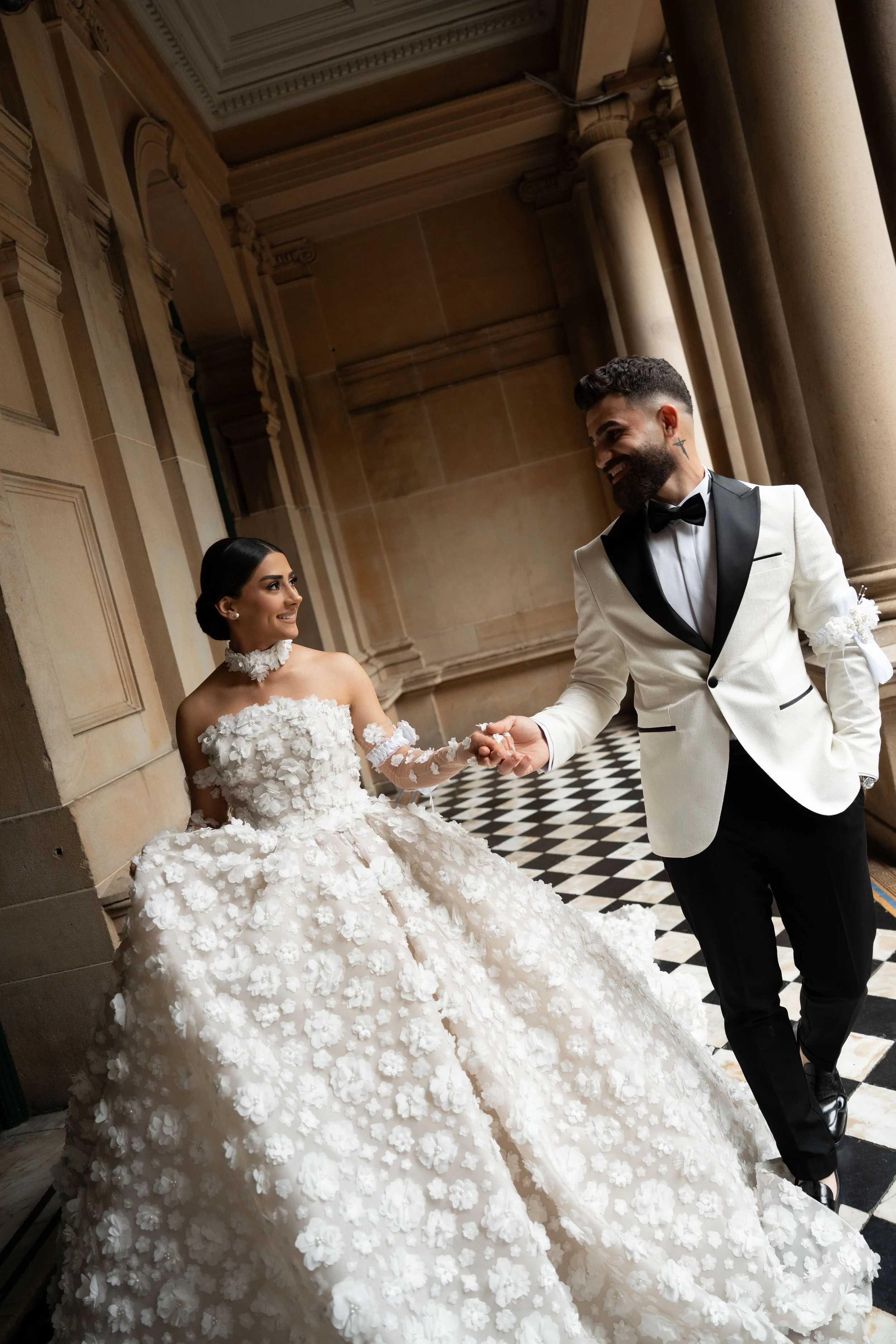 A bride and groom on their wedding day, holding hands and smiling at each other, indoors with elegant architectural details.