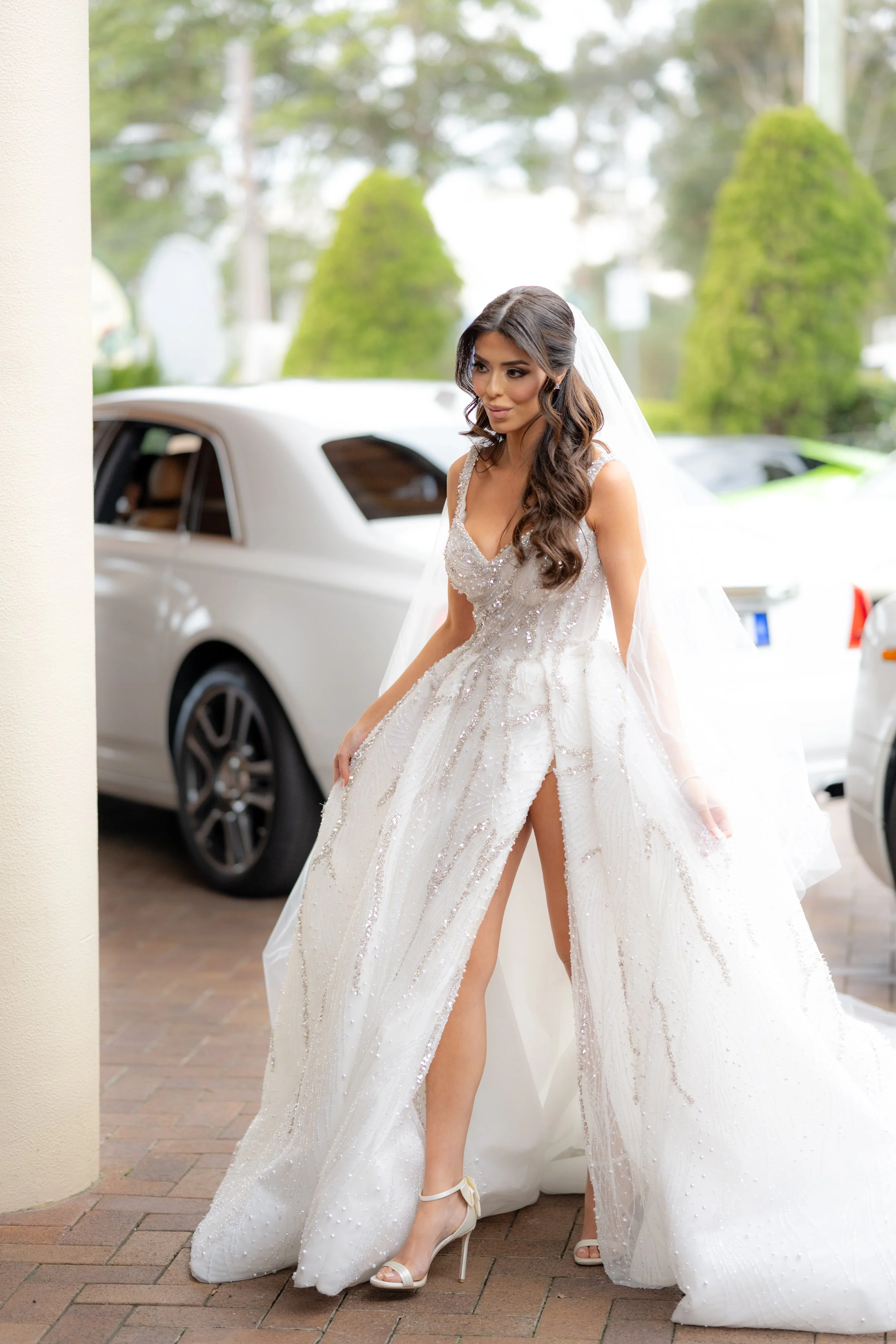 A woman in a white wedding gown with intricate beadwork and a high slit, standing next to a luxury car outdoors, with trees in the background.