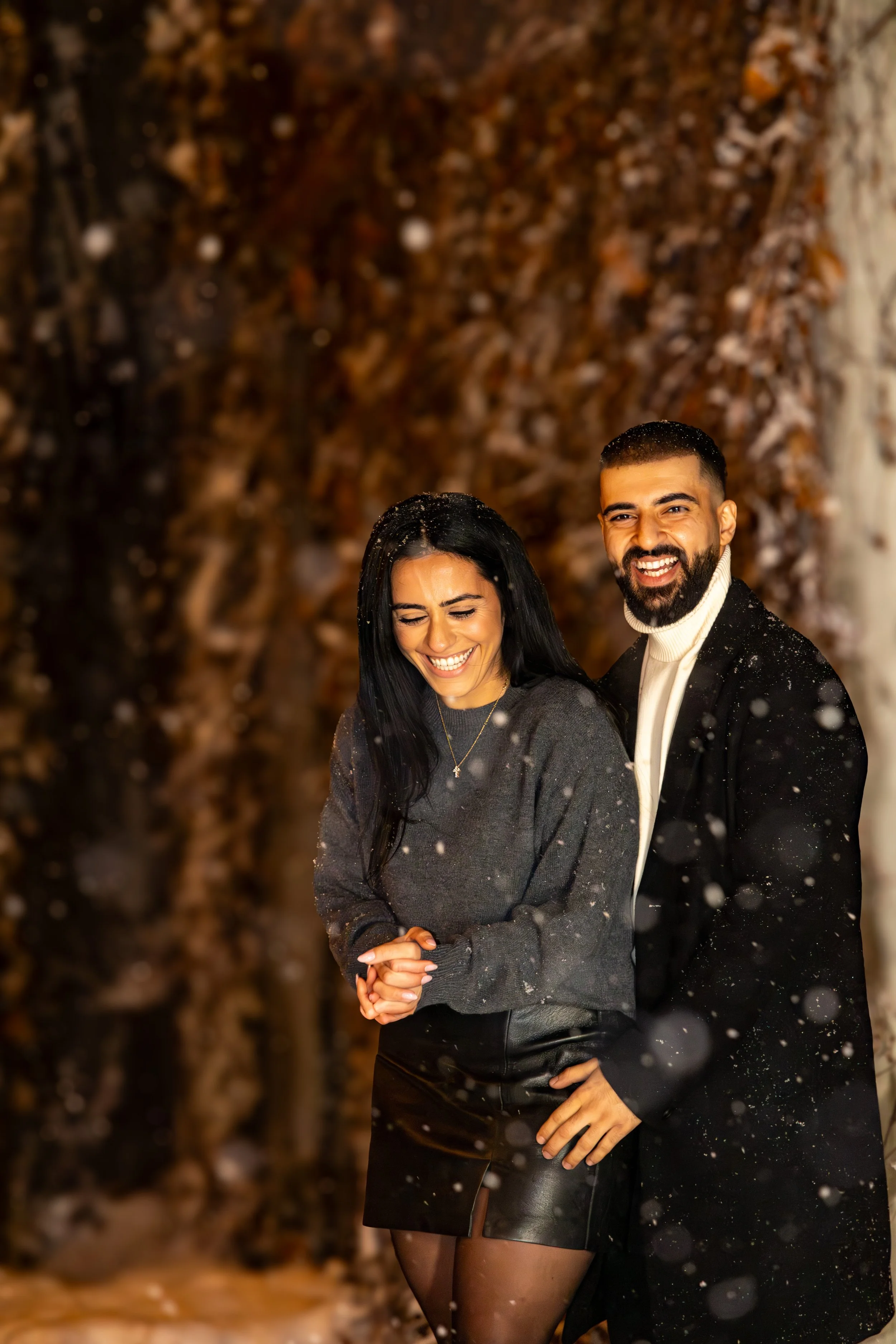A smiling couple standing outdoors in the snow, with a background of trees with brown leaves.