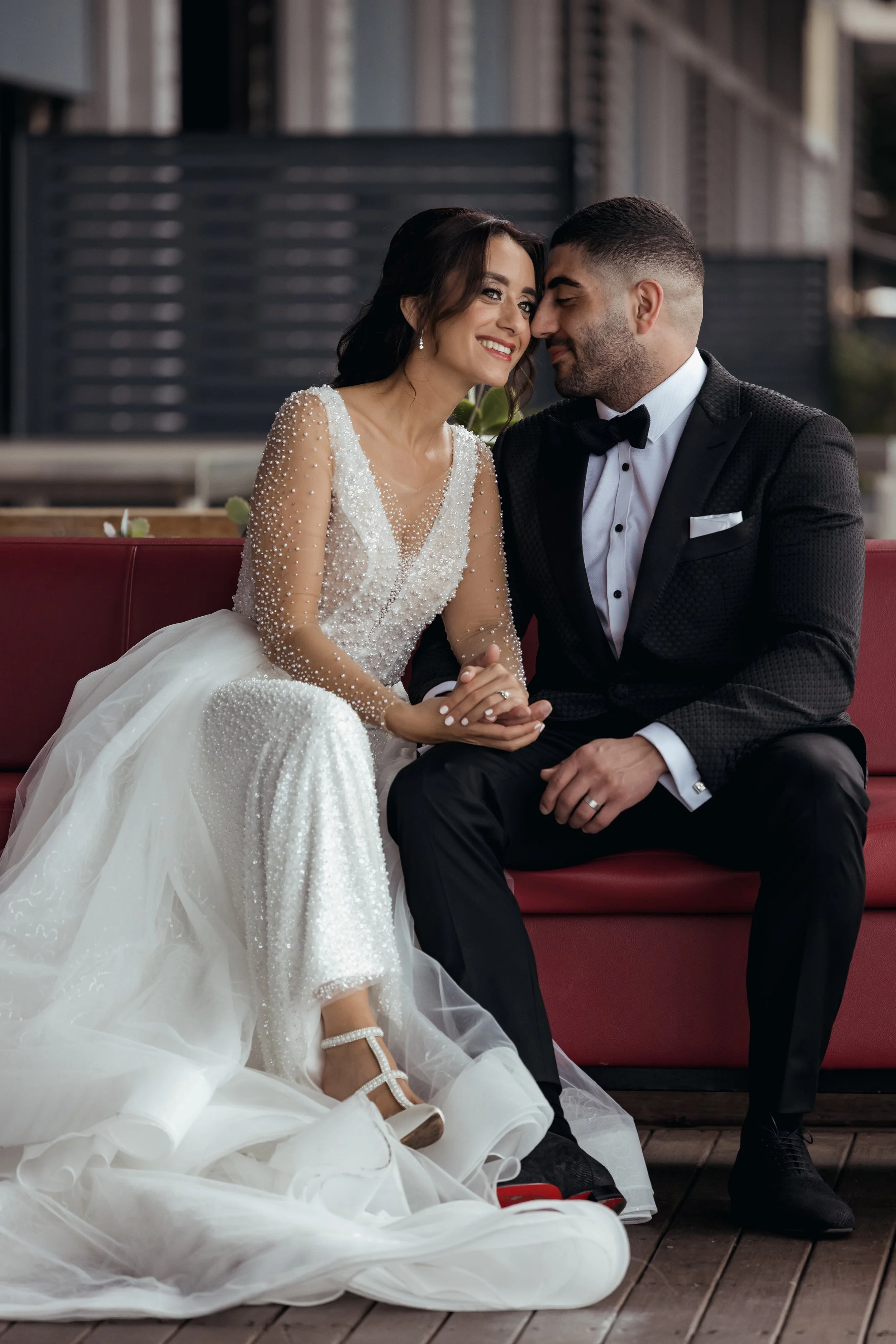A bride and groom sitting close together, smiling affectionately. The bride wears a white wedding gown with pearl embellishments and white high heels, while the groom wears a black tuxedo with a bow tie, white shirt, and black dress shoes. They are seated outdoors on a red bench with a modern building in the background.