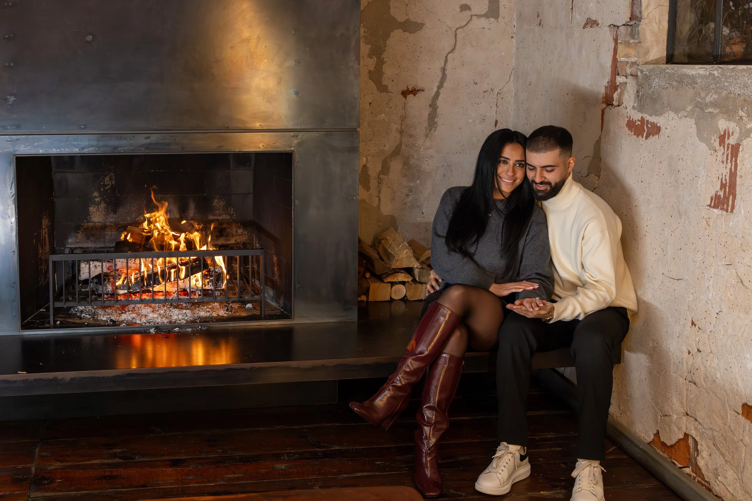 A smiling woman and man sitting next to each other by a fireplace, looking at a phone, with a pile of firewood beside them.