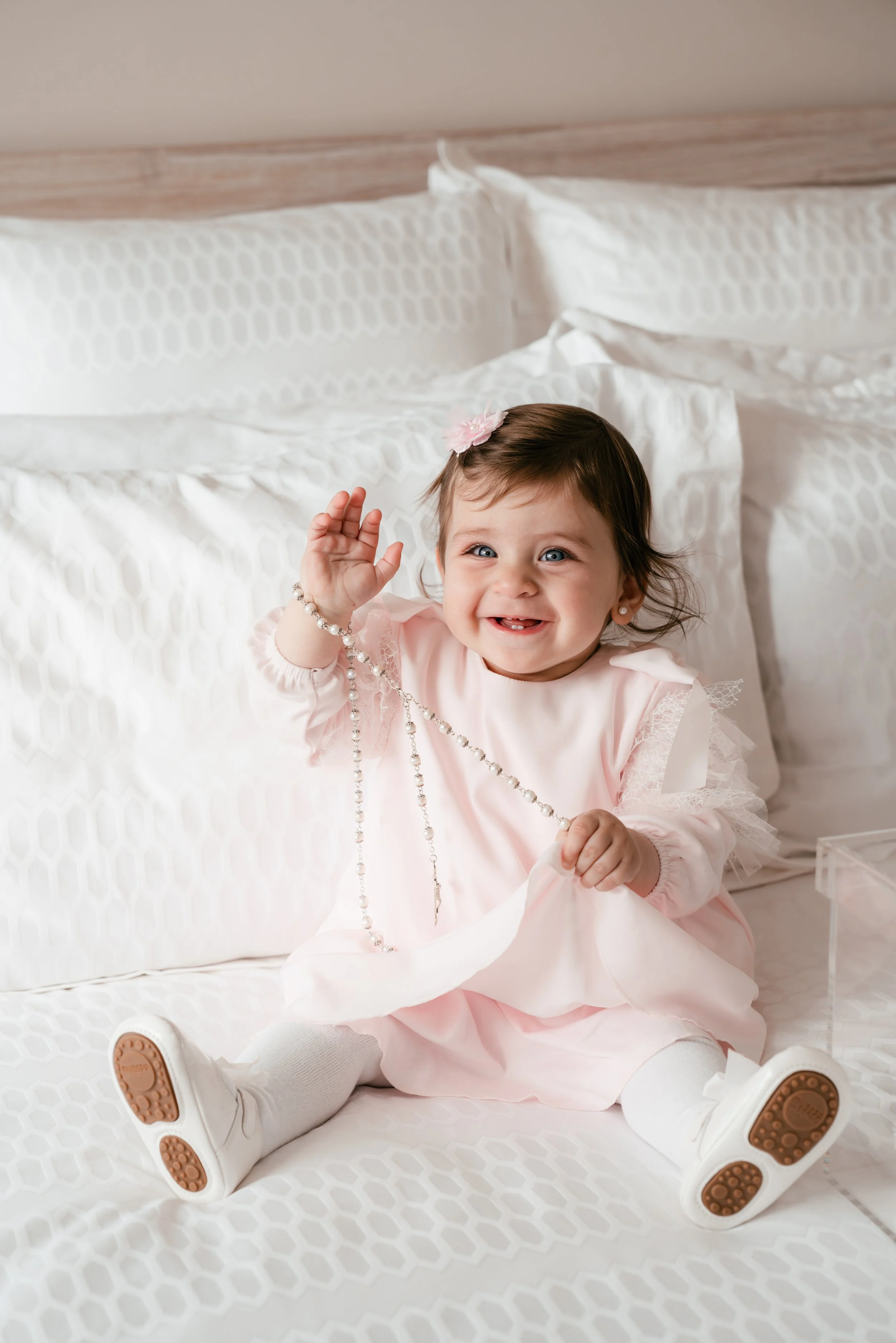 A smiling toddler girl with dark hair, blue eyes, and a pink flower hair clip, sitting on a white bed with geometric patterned bedding, holding a pearl necklace.