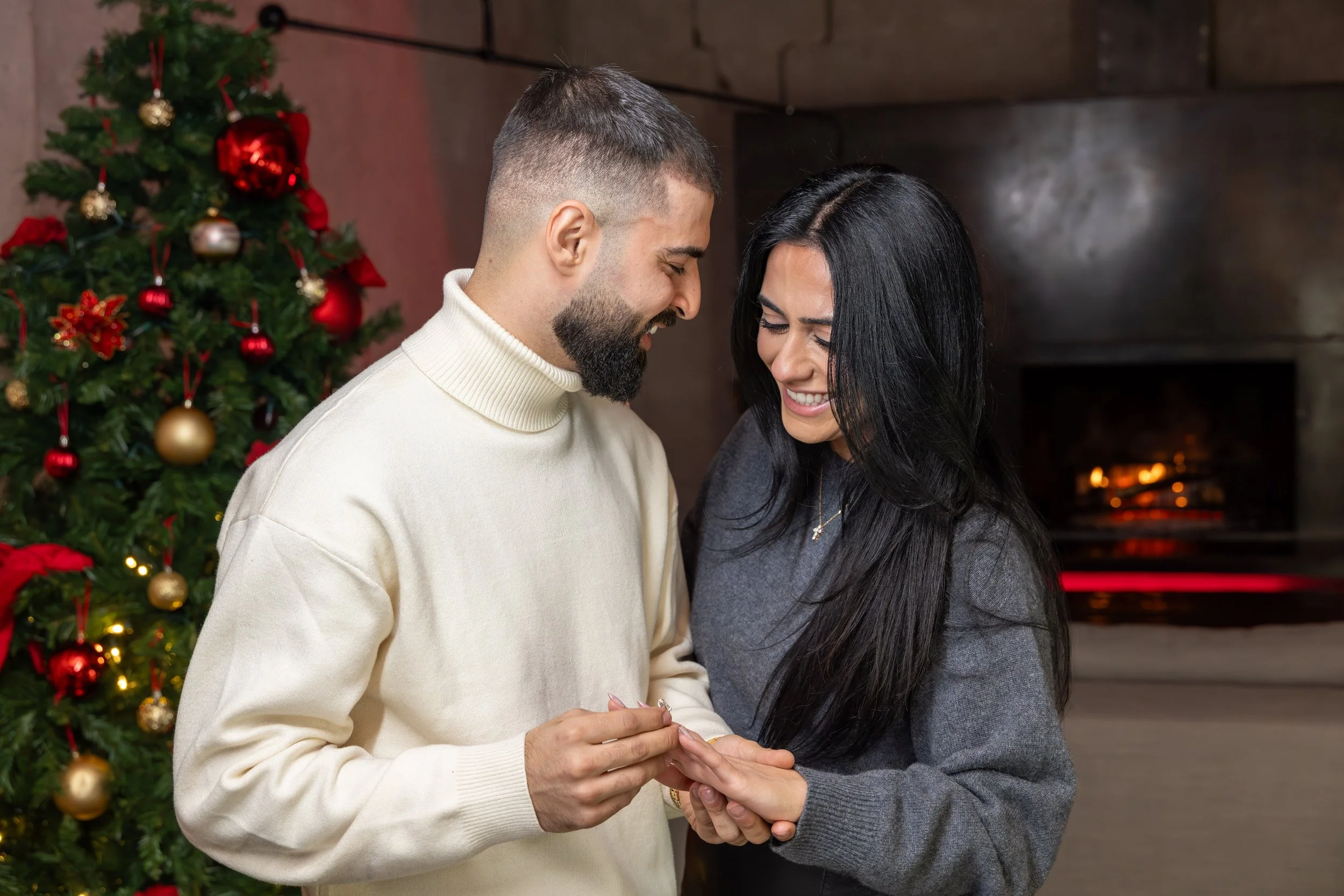 A couple exchanging rings near a decorated Christmas tree with a fireplace in the background.