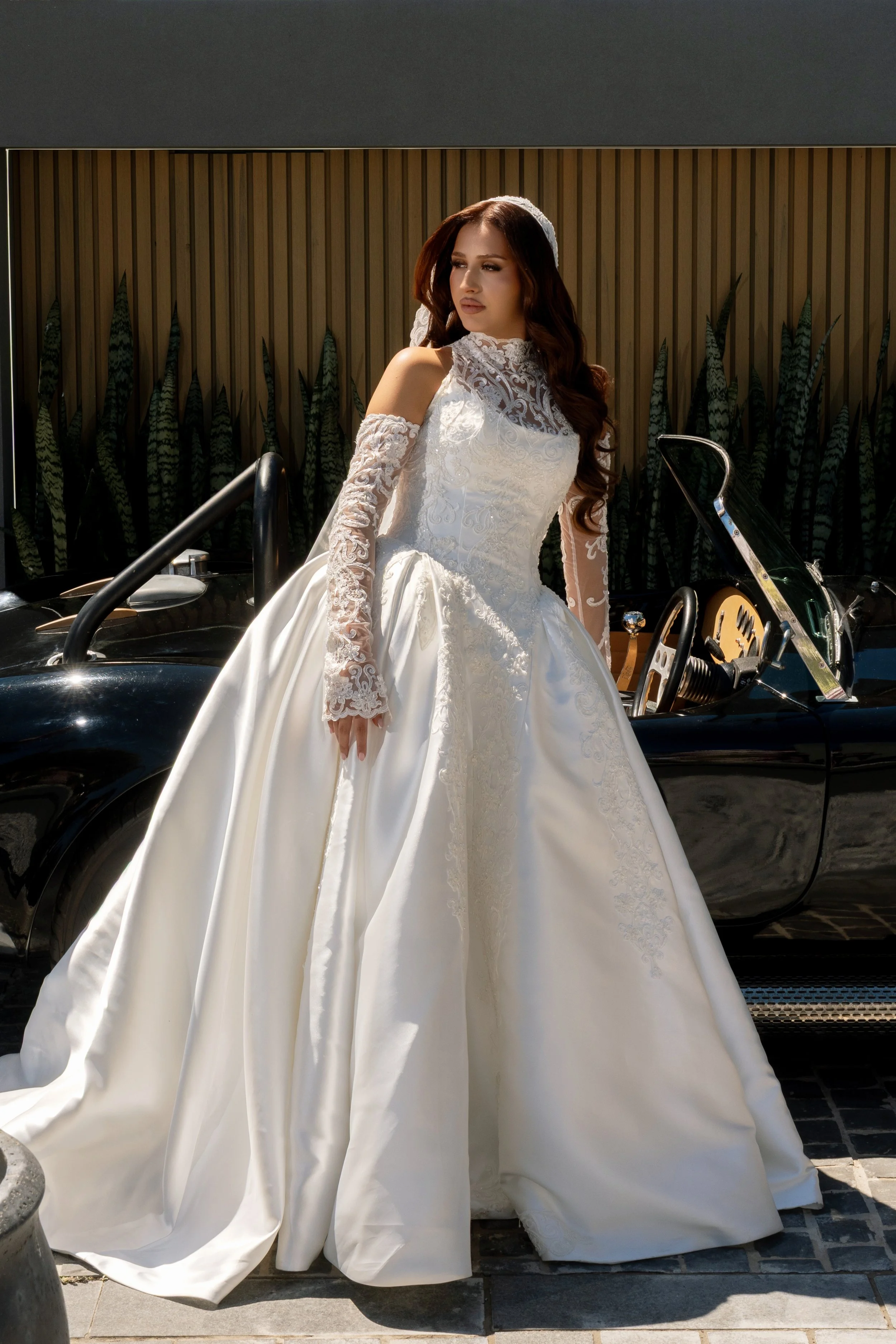 A woman in a white wedding gown standing beside a black vintage car with an open door, background featuring a wooden wall and tall plants.