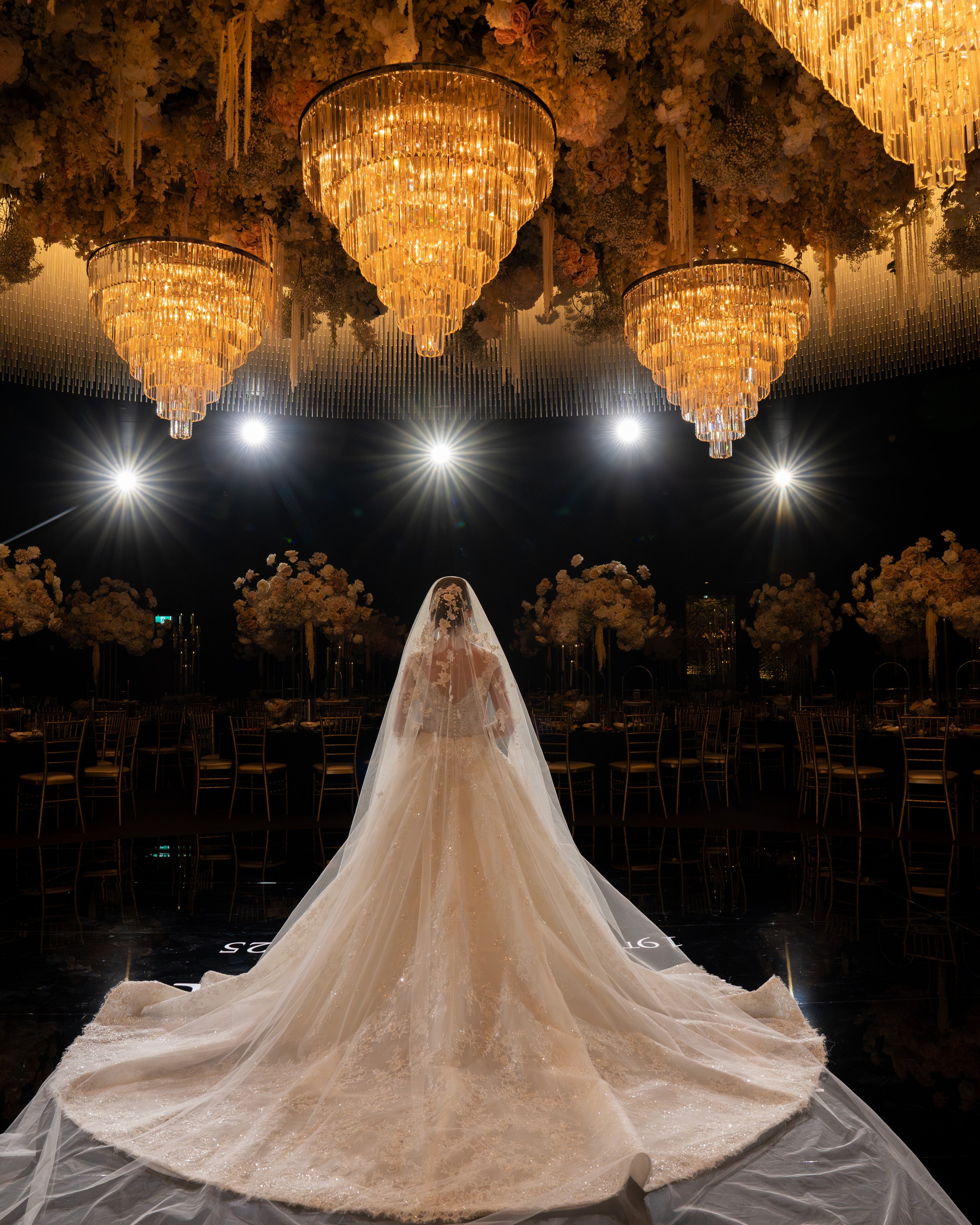 A bride in a wedding gown with a long, flowing train and veil, standing under ornate chandeliers in a decorated event hall with floral arrangements and dining tables.