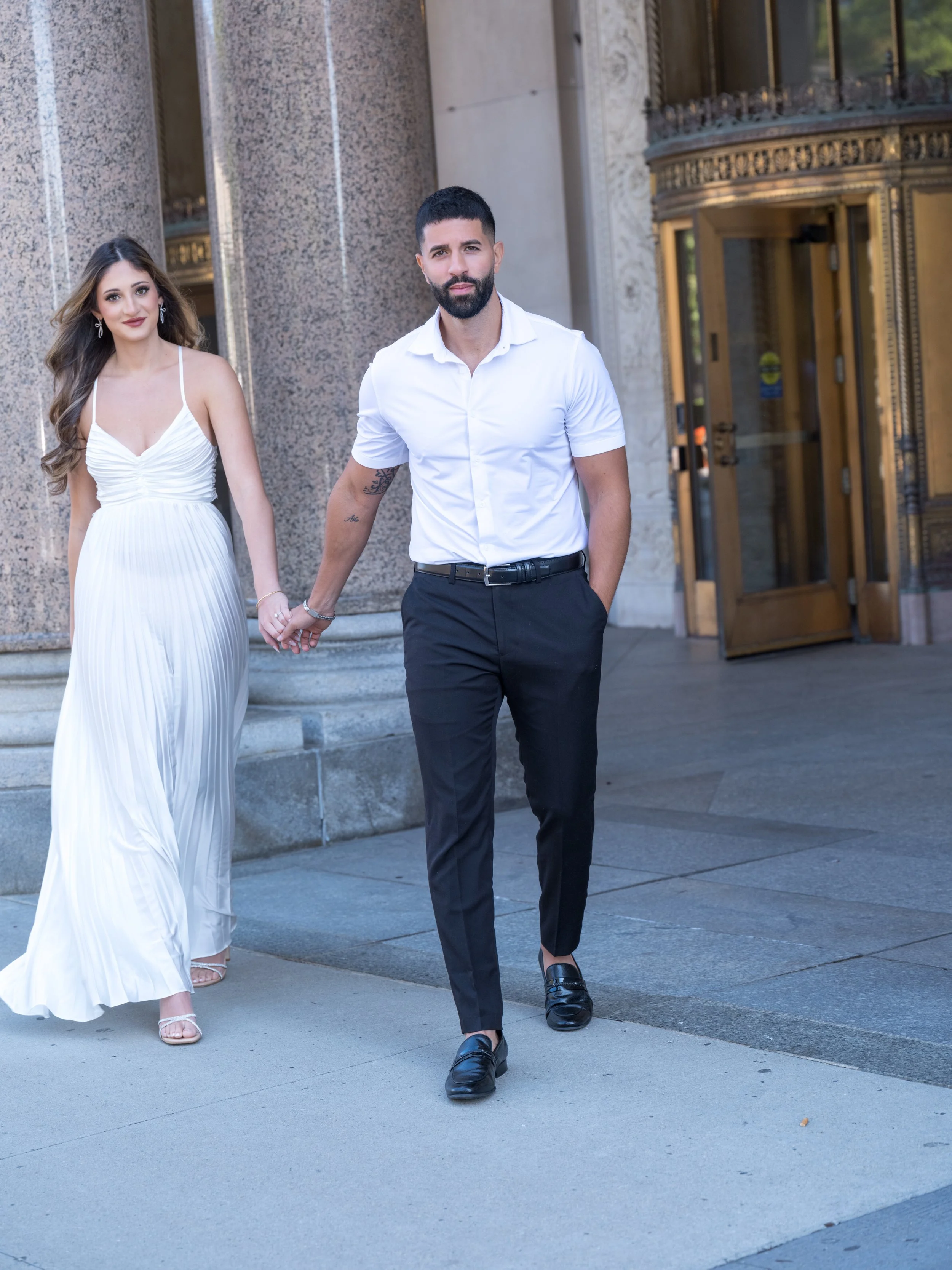 A young man and woman holding hands while walking outside a building. The woman is wearing a white strapless dress with pleats and high heels, and the man is dressed in a white short-sleeved shirt, dark pants, and black loafers.