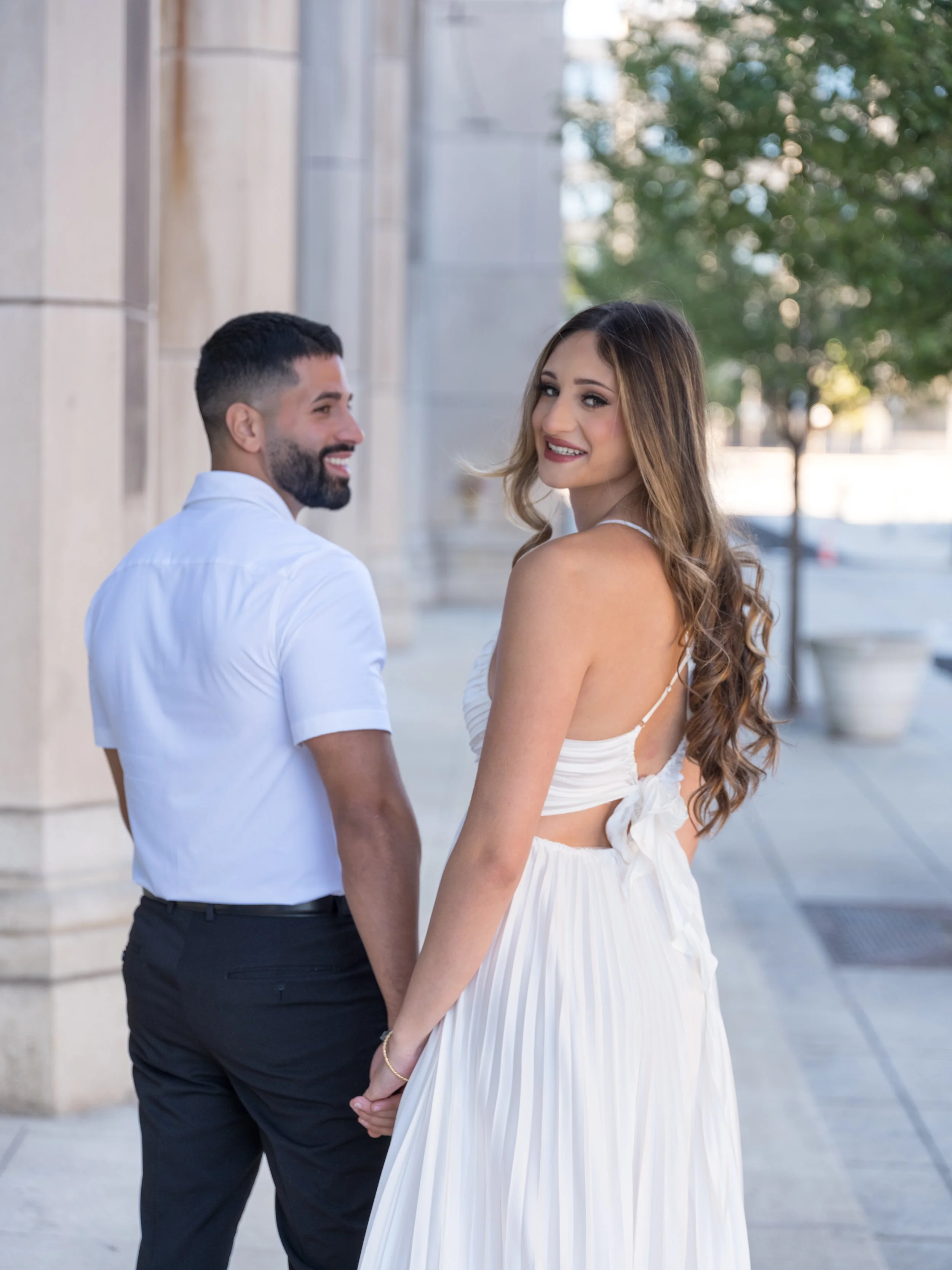 A happy couple holding hands on city sidewalk, smiling at each other, woman in white dress and man in white shirt and black pants.