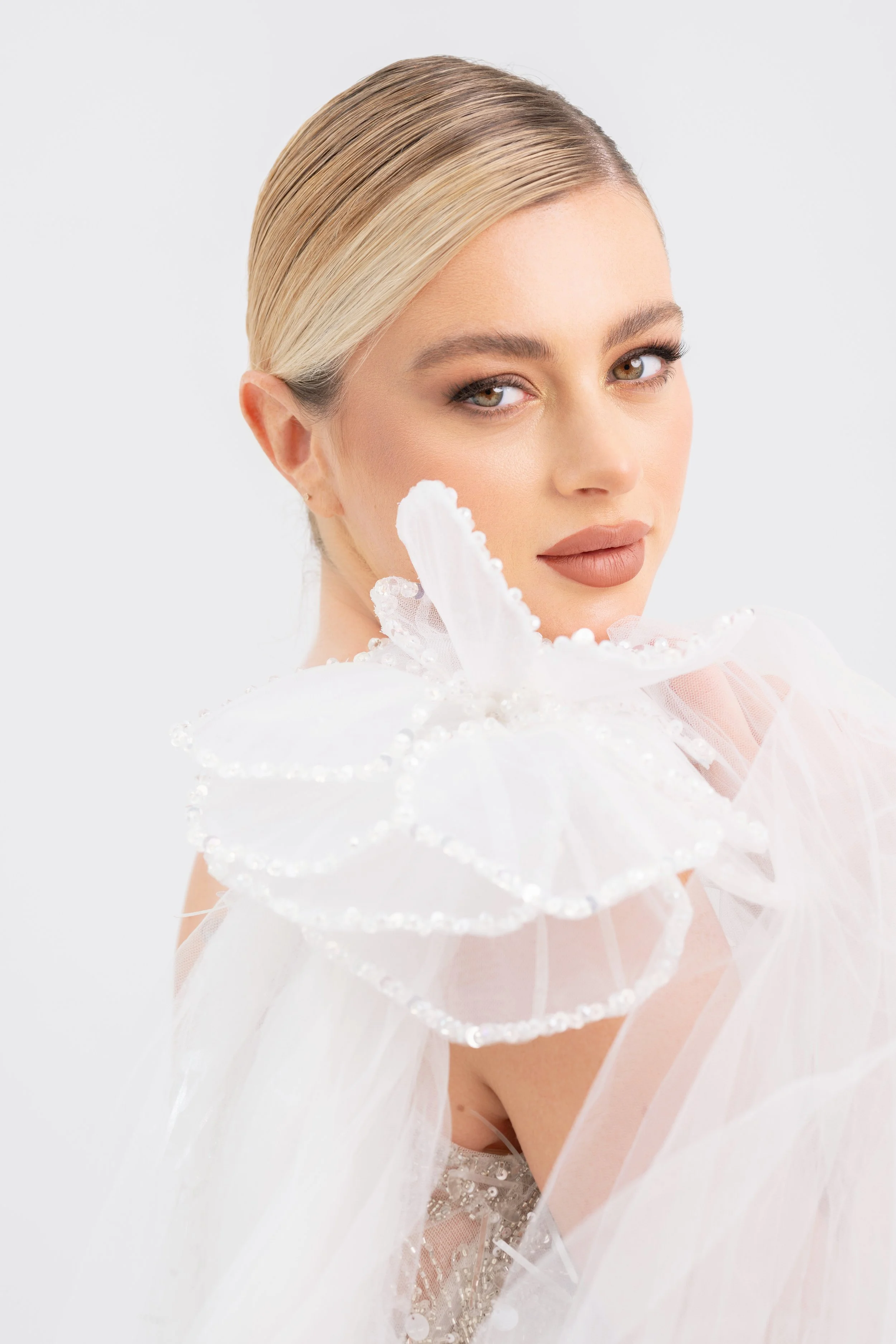 A woman with blonde hair styled in a sleek side part, wearing makeup, with a neutral expression. She is holding a white, sheer, ruffled fabric with pearl embellishments near her shoulder, against a plain white background.