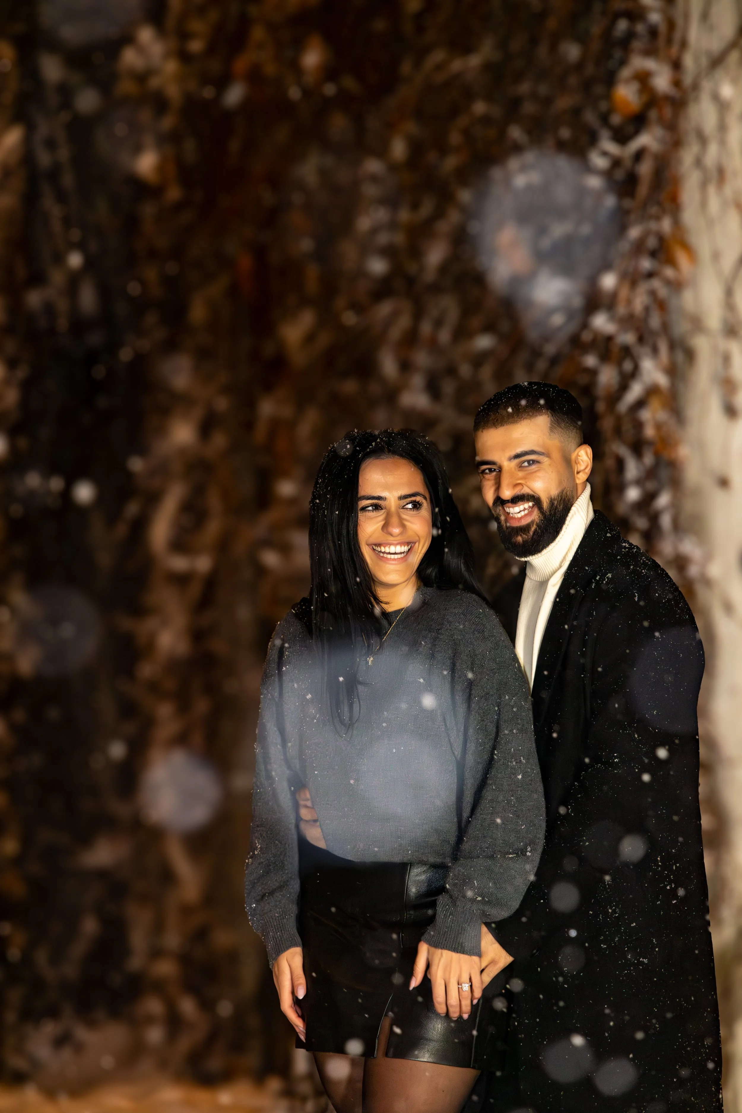 A smiling woman and man standing close together outdoors in the snow at night, with a blurred background of tree branches.