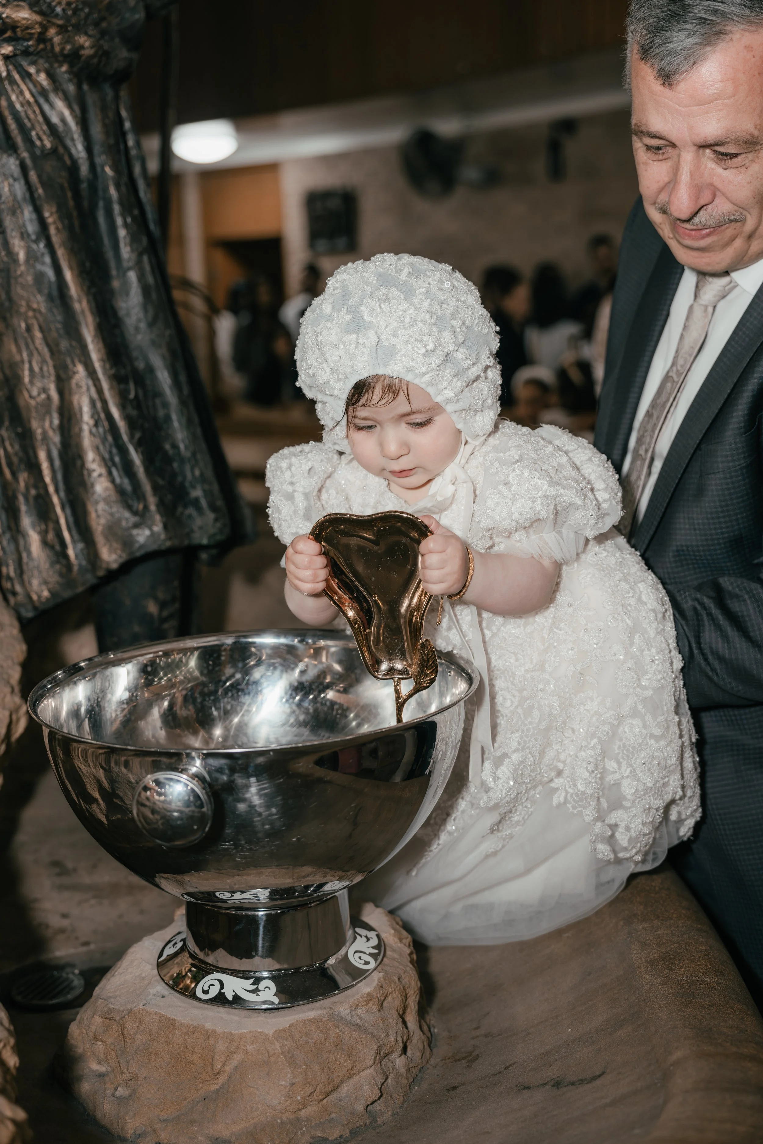 Child dressed in white with lace and floral details, wearing a matching bonnet, pouring honey into a large silver bowl held by an adult man in a dark suit at a ceremonial event.