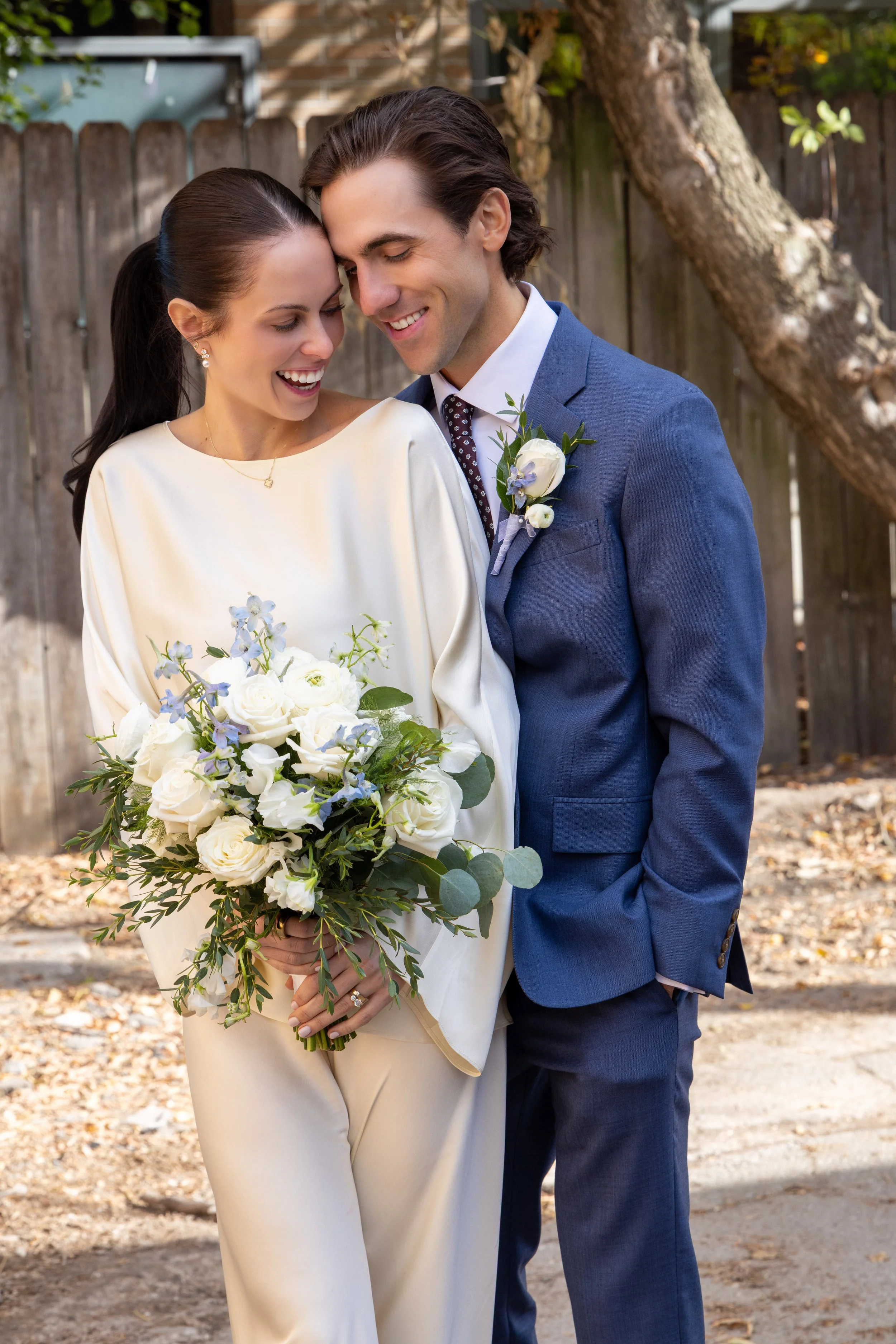 A couple dressed in wedding attire, smiling and leaning their heads together outdoors, with a wooden fence and tree in the background. The woman holds a bouquet of white and purple flowers, and the man wears a blue suit with a boutonniere.