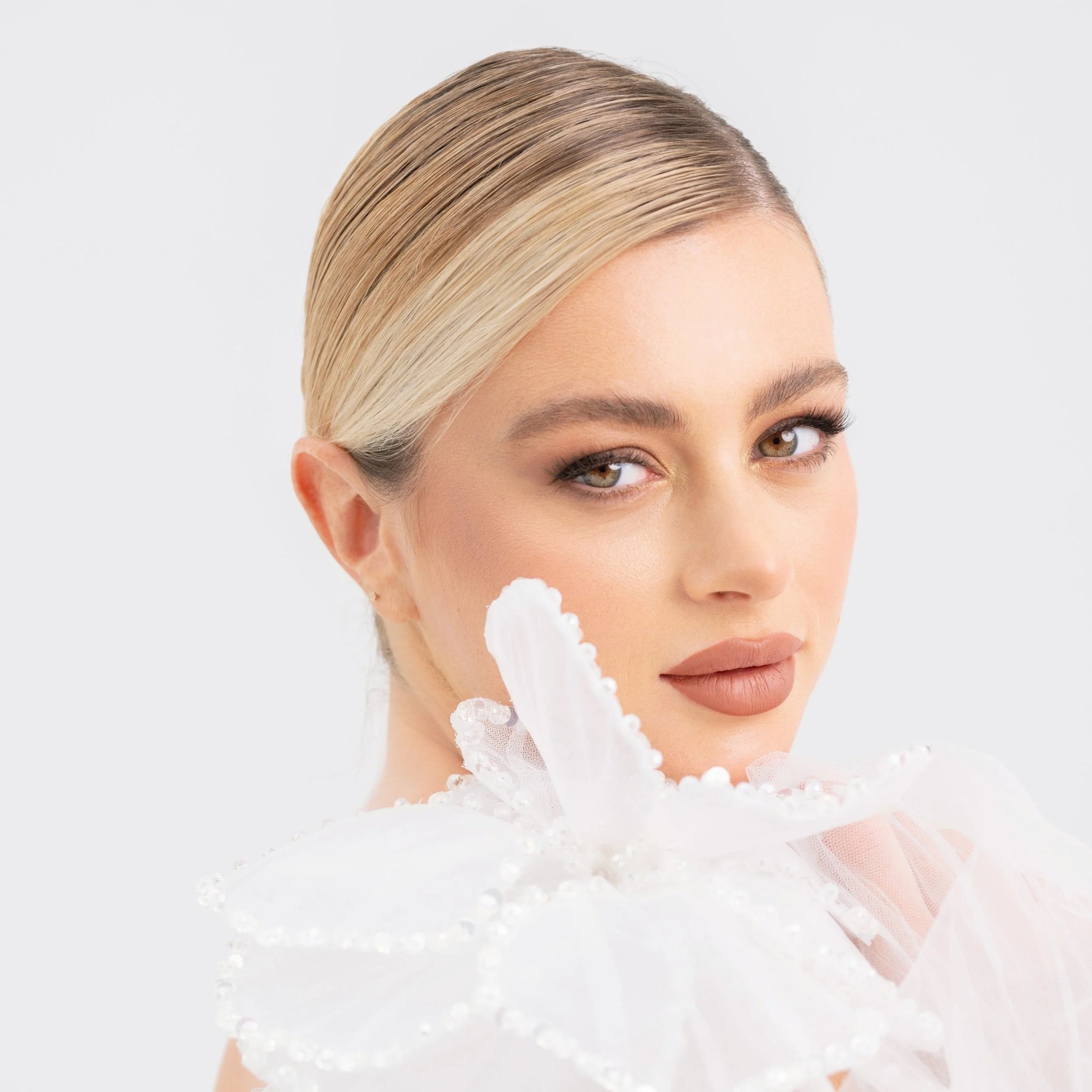 A woman with blonde hair styled back, wearing neutral makeup, and a white dress with large, decorative white flower details with pearl accents.