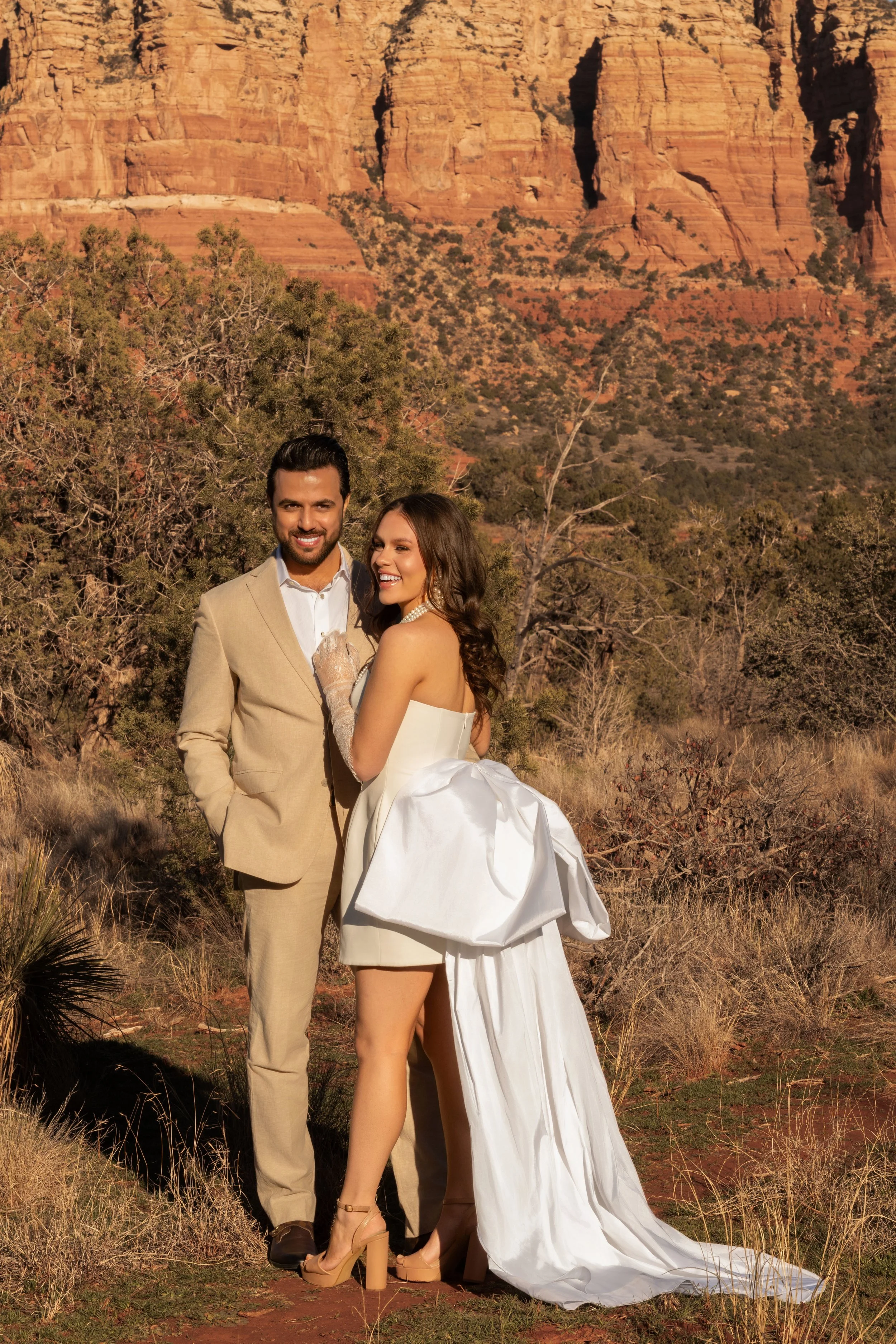 A couple dressed in wedding attire, standing in a desert landscape with red rock formations and sparse vegetation.