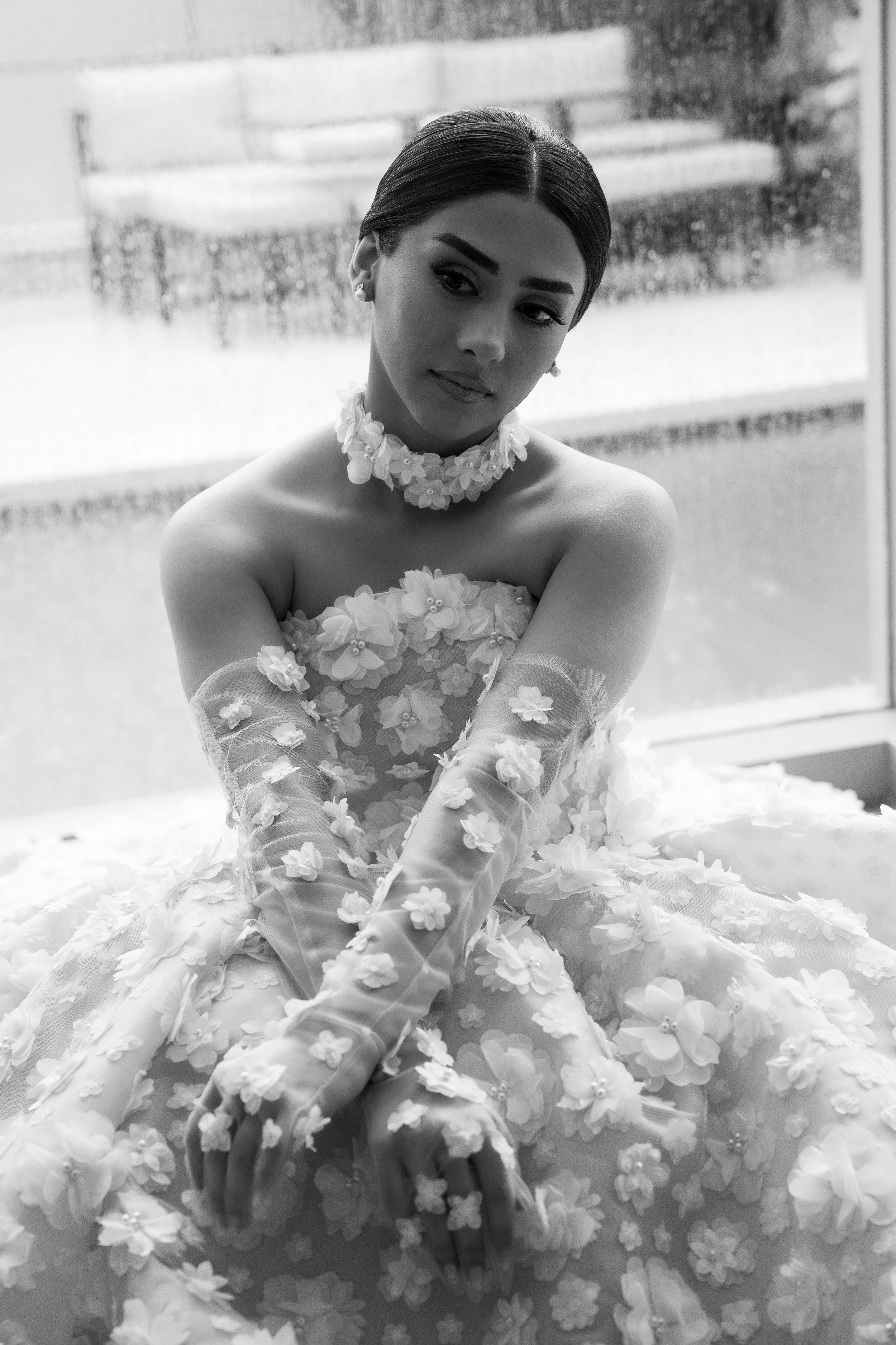 Black-and-white photo of a woman in an ornate, floral gown with long gloves, sitting by a window on a rainy day.