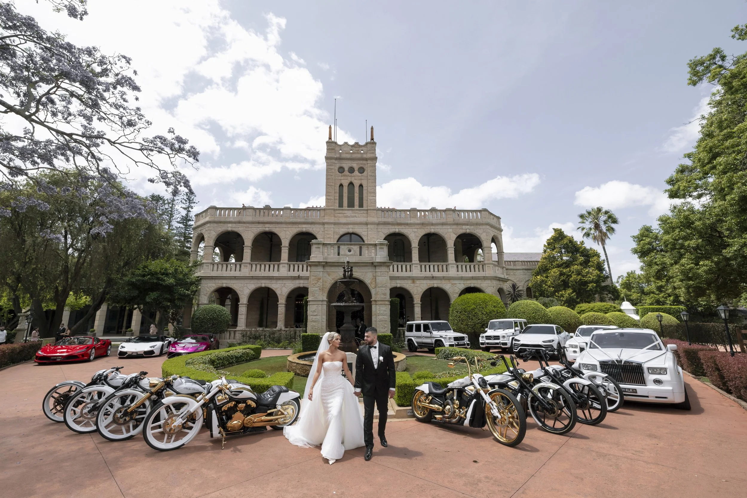Bride and groom walking hand in hand in front of a luxurious mansion with cars and motorcycles parked nearby, lush greenery in the background.